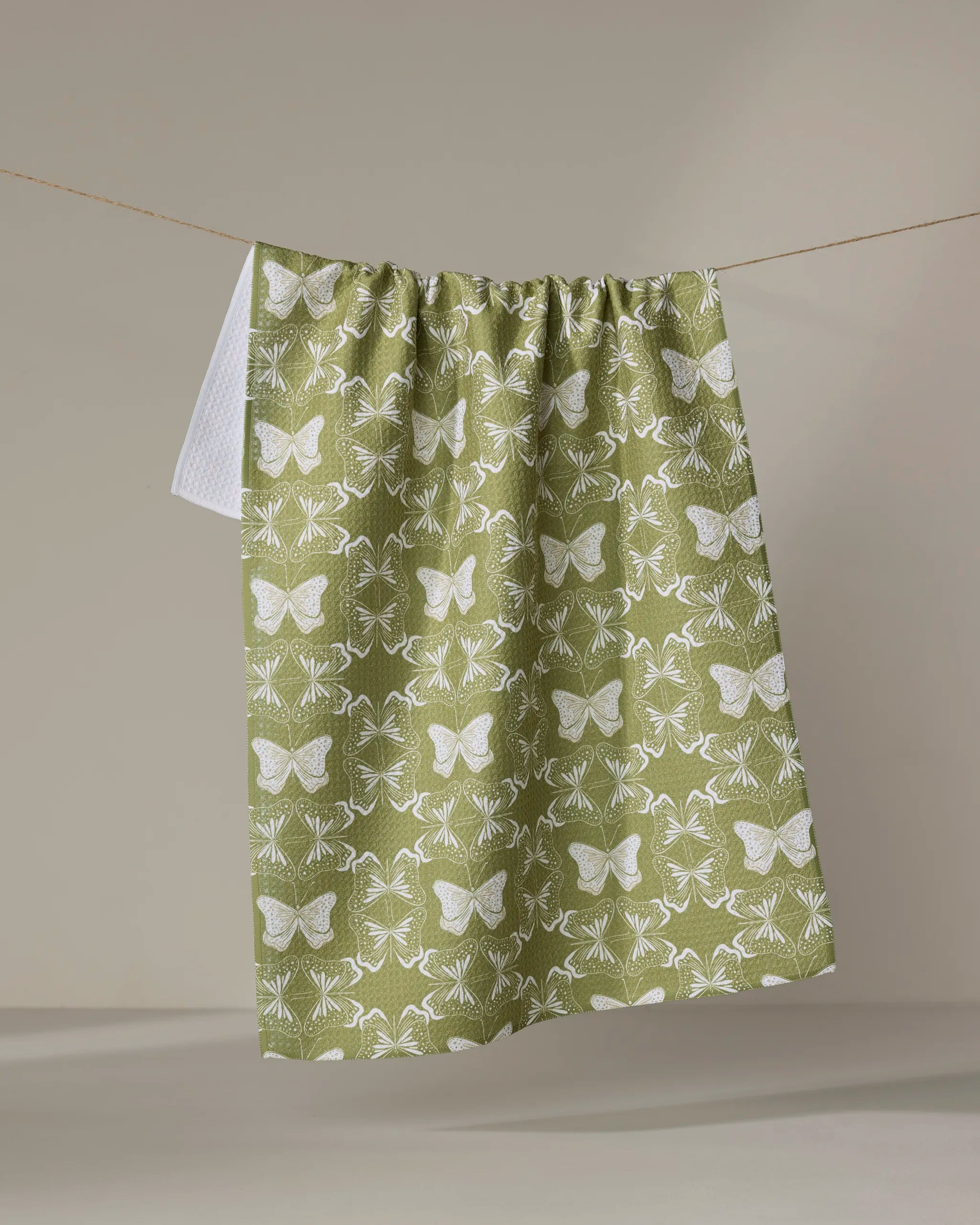 The "Geometric Butterflies" green tea towel by Second Nature Studio, made from recycled materials, features a white butterfly and floral pattern and hangs over a clothesline against a neutral background.