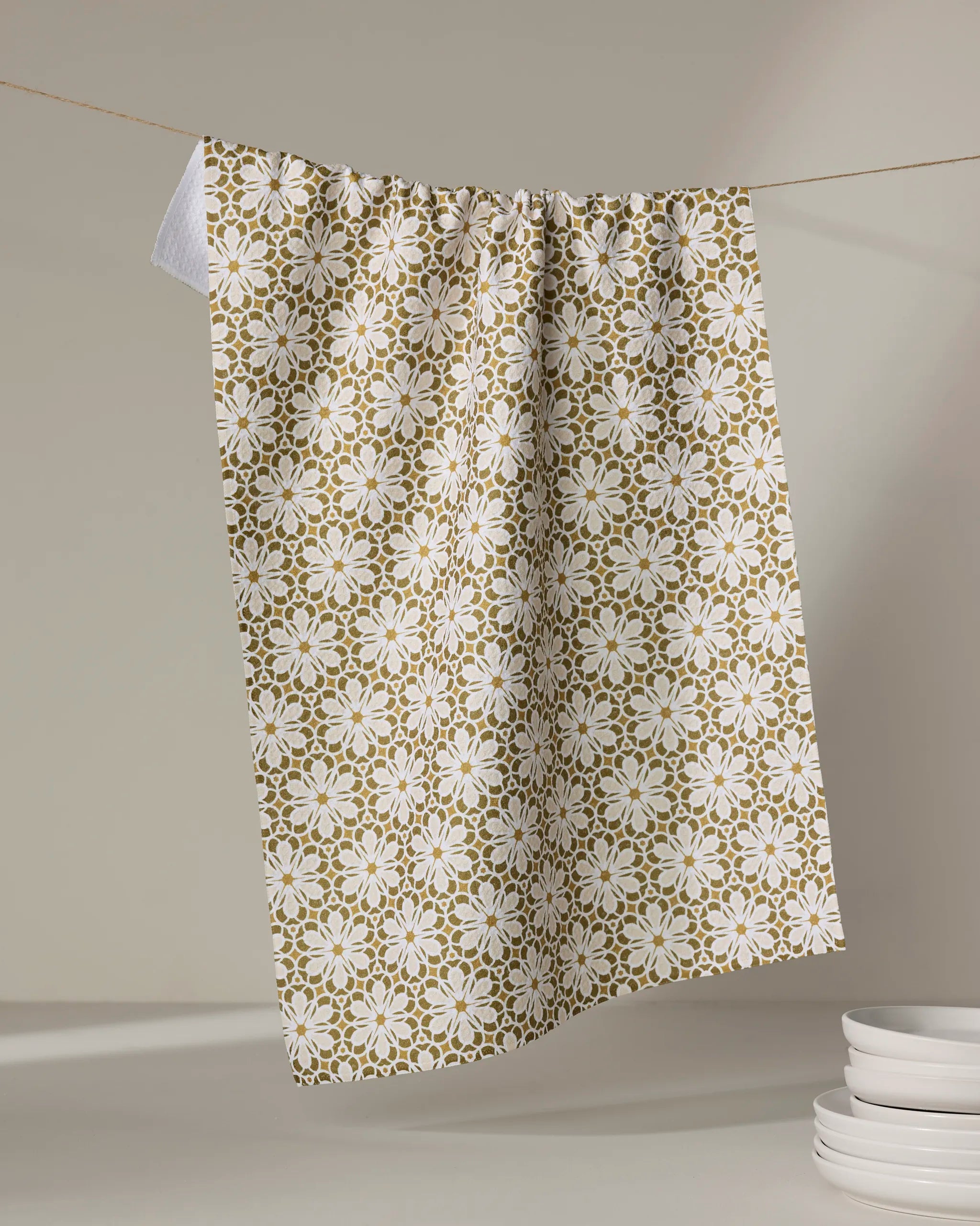 The Geometric Flower tea towel by Marta Harvey, featuring a green and white floral pattern, hangs on a string against a neutral background with a stack of white plates in the lower right corner.