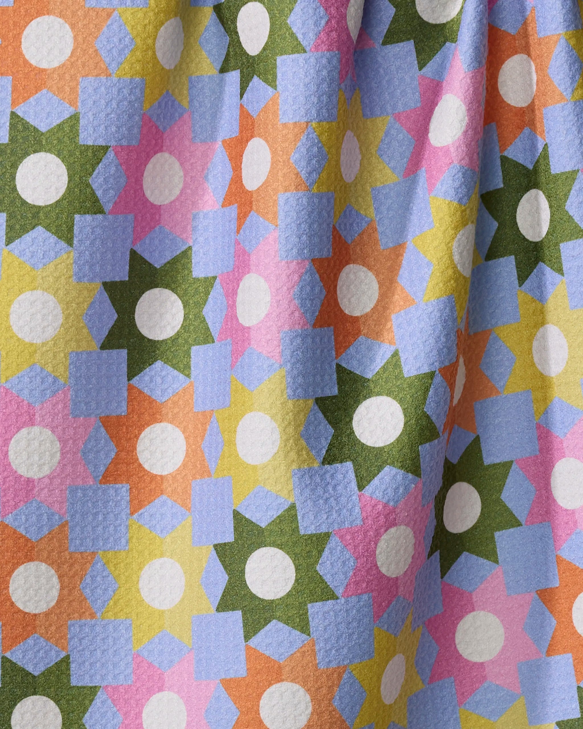A close-up of Megan Roy’s Geometric Garden fabric, made from recycled materials, shows textured, draped material with blue squares, white circles, green stars, and orange, pink, and yellow geometric shapes in a repeating pattern.