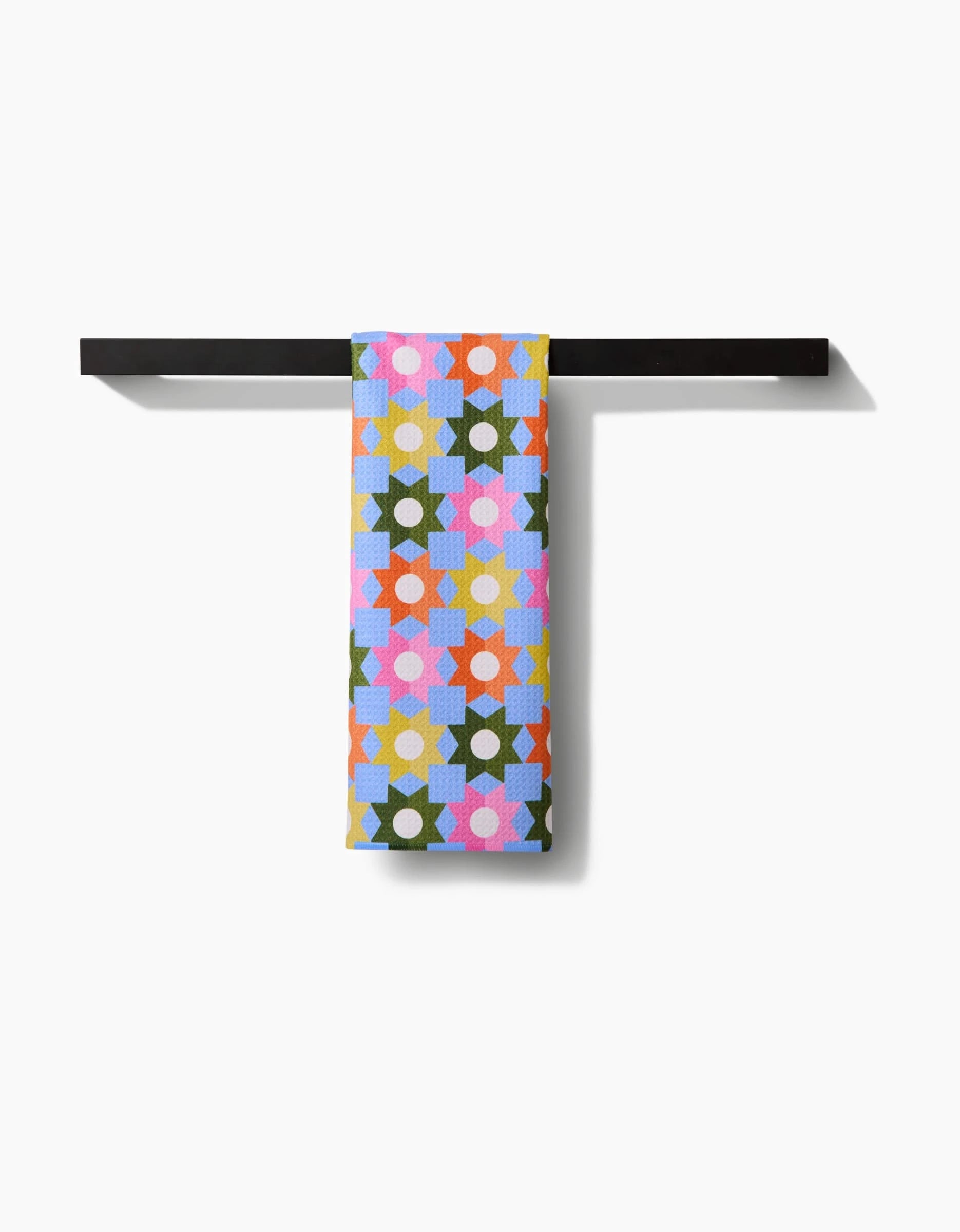 The Megan Roy Geometric Garden towel features a vibrant geometric floral pattern and hangs on a black bar. Made from ultra-absorbent Geoweave fabric, it adds lively color against a white background.