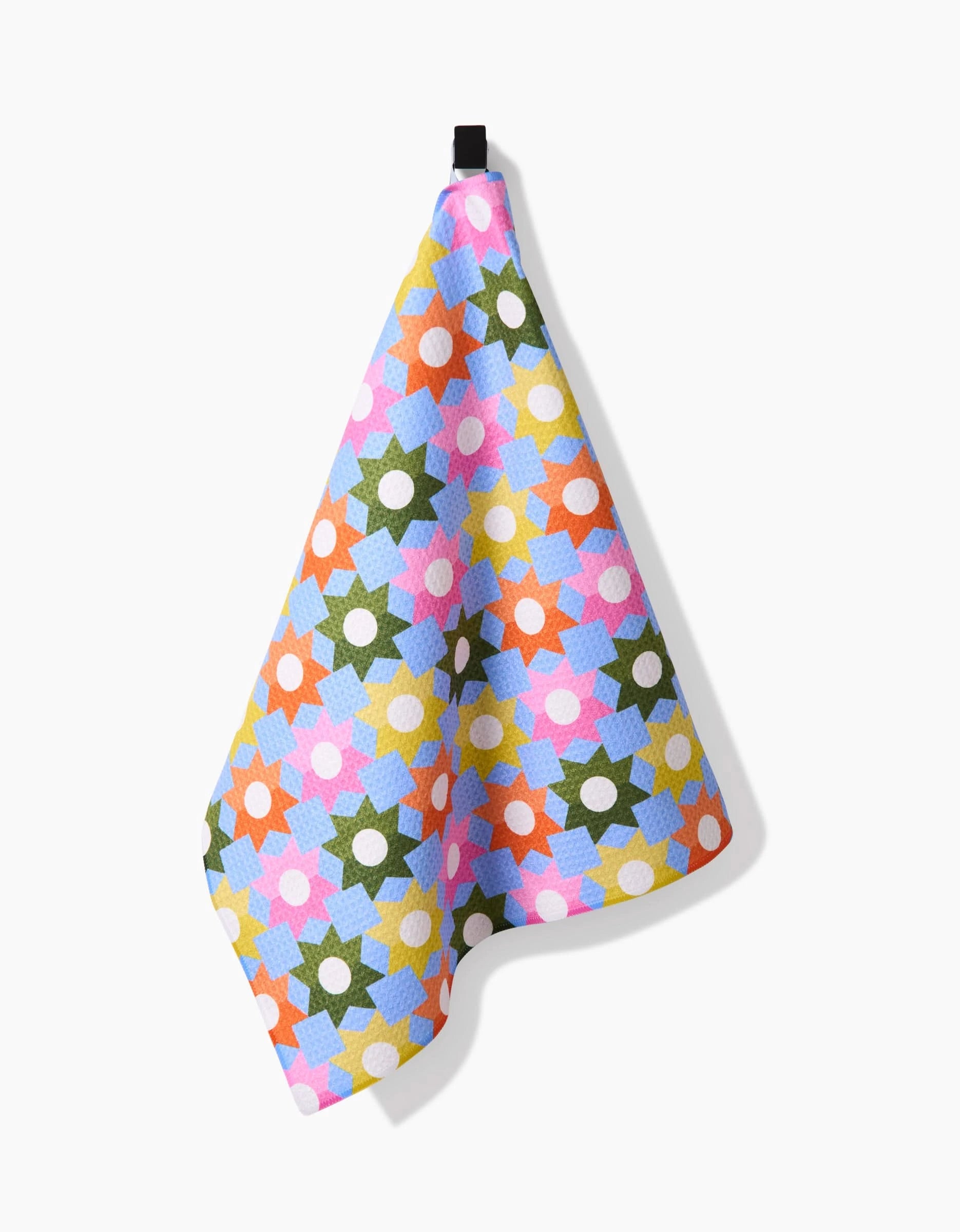 The Megan Roy “Geometric Garden” tea towel features a geometric floral pattern in green, blue, pink, red, and yellow on ultra-absorbent Geoweave fabric, shown hanging from a black loop against a white background.