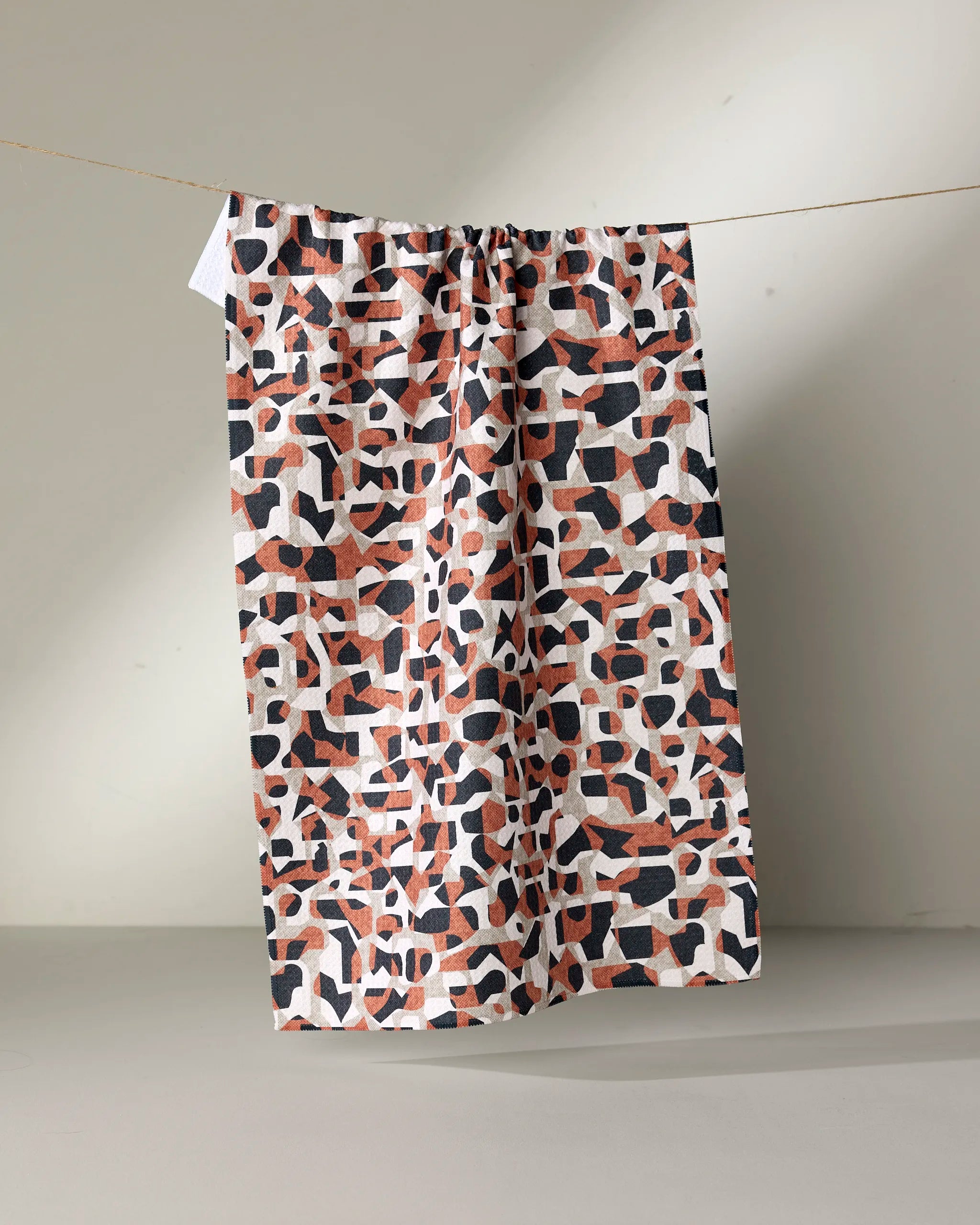 The Geometric Leopard tea towel by Kris Ruff features abstract shapes in black, white, and rust. Made from recycled materials, this eco-friendly and stylish piece hangs on a thin line against a neutral background.