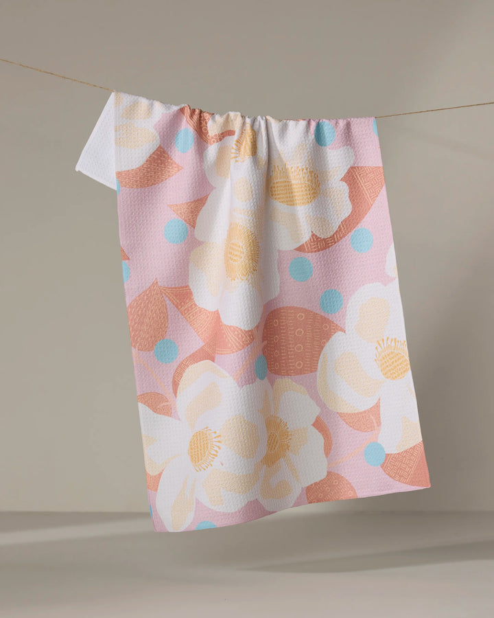 The "Georgia Rose" tea towel by Julianne Haness, featuring large white and yellow flowers, peach motifs, and blue polka dots in pastel hues, hangs on a clothesline against a simple beige background.