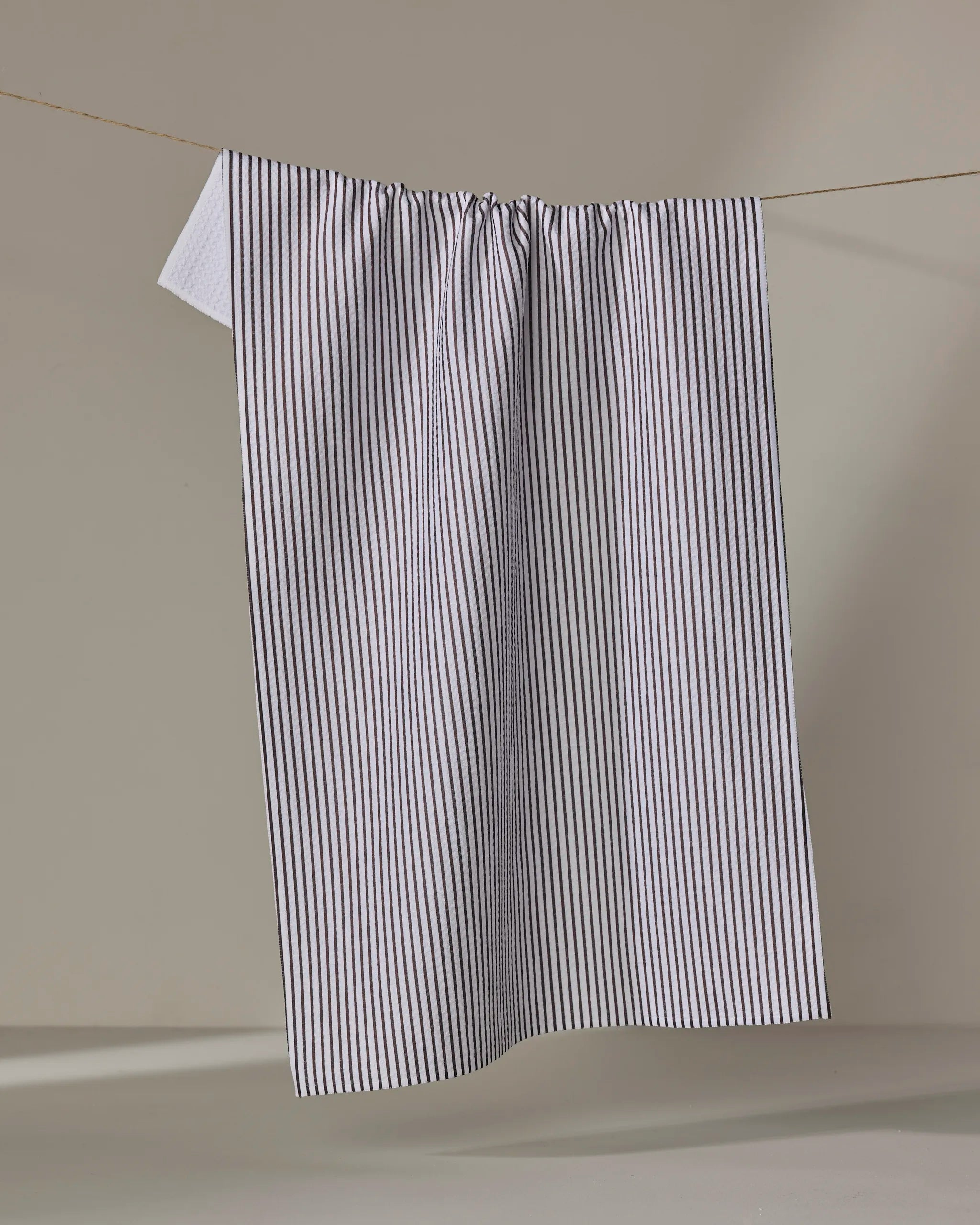 The Buy Guide's Get In Line - Chocolate towel, featuring thin black vertical stripes on a white ultra-absorbent fabric, hangs from a clothesline against a plain, light background.