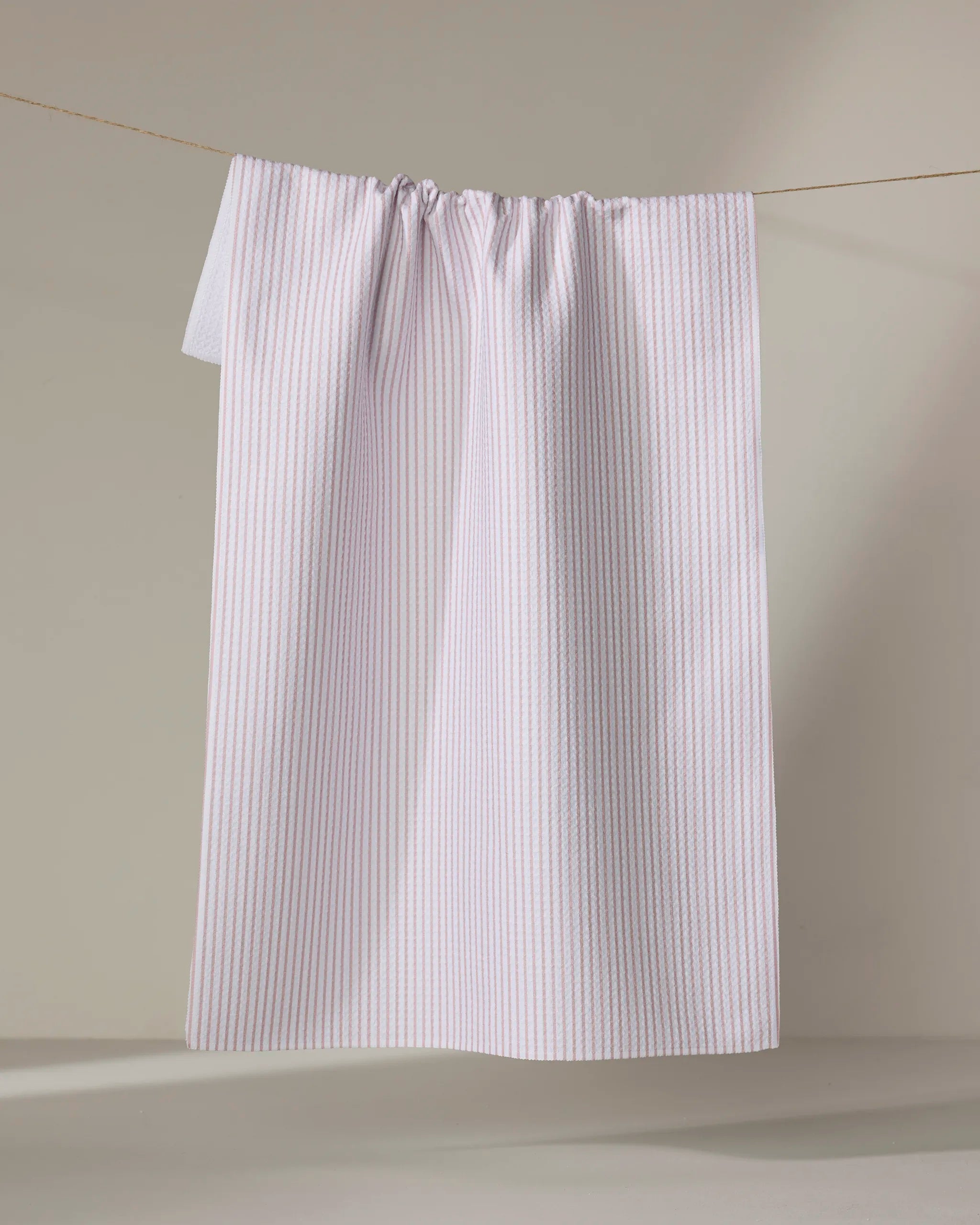 The Buy Guide's Get In Line - Dusty Rose towel, featuring thin vertical pink stripes on white, hangs over a clothesline against a neutral background.