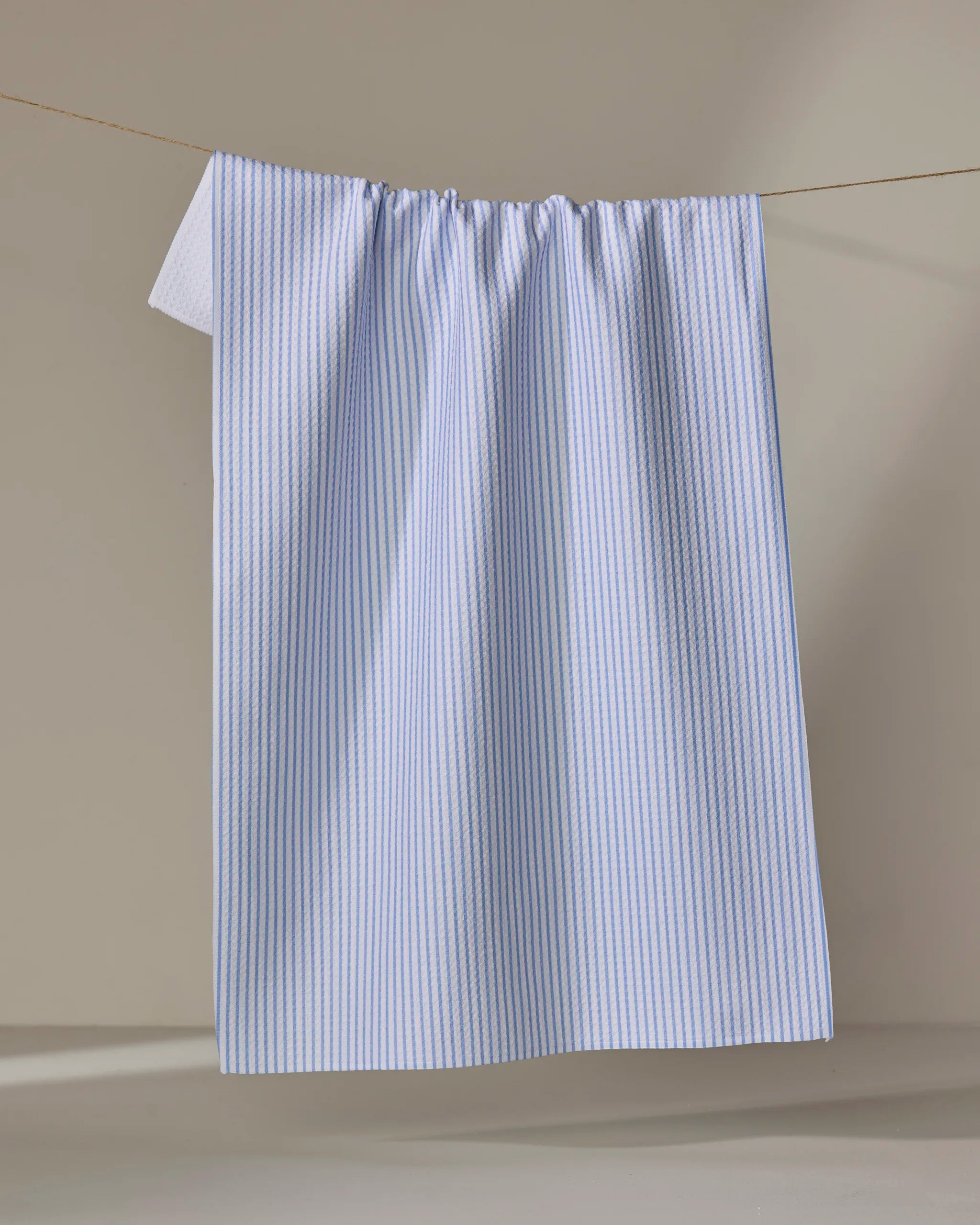 The Buy Guide’s Get In Line - French Blue tea towel, featuring light blue and white stripes and made from recycled materials, hangs neatly on a clothesline against a neutral background.