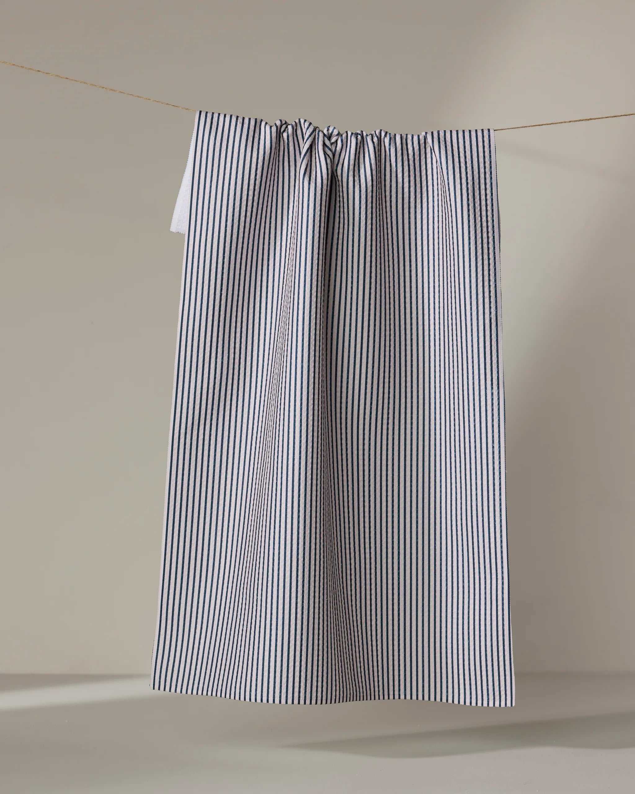 The Buy Guide's "Get In Line - Navy" tea towel, featuring thin vertical navy stripes on white, hangs neatly on a clothesline against a neutral background.