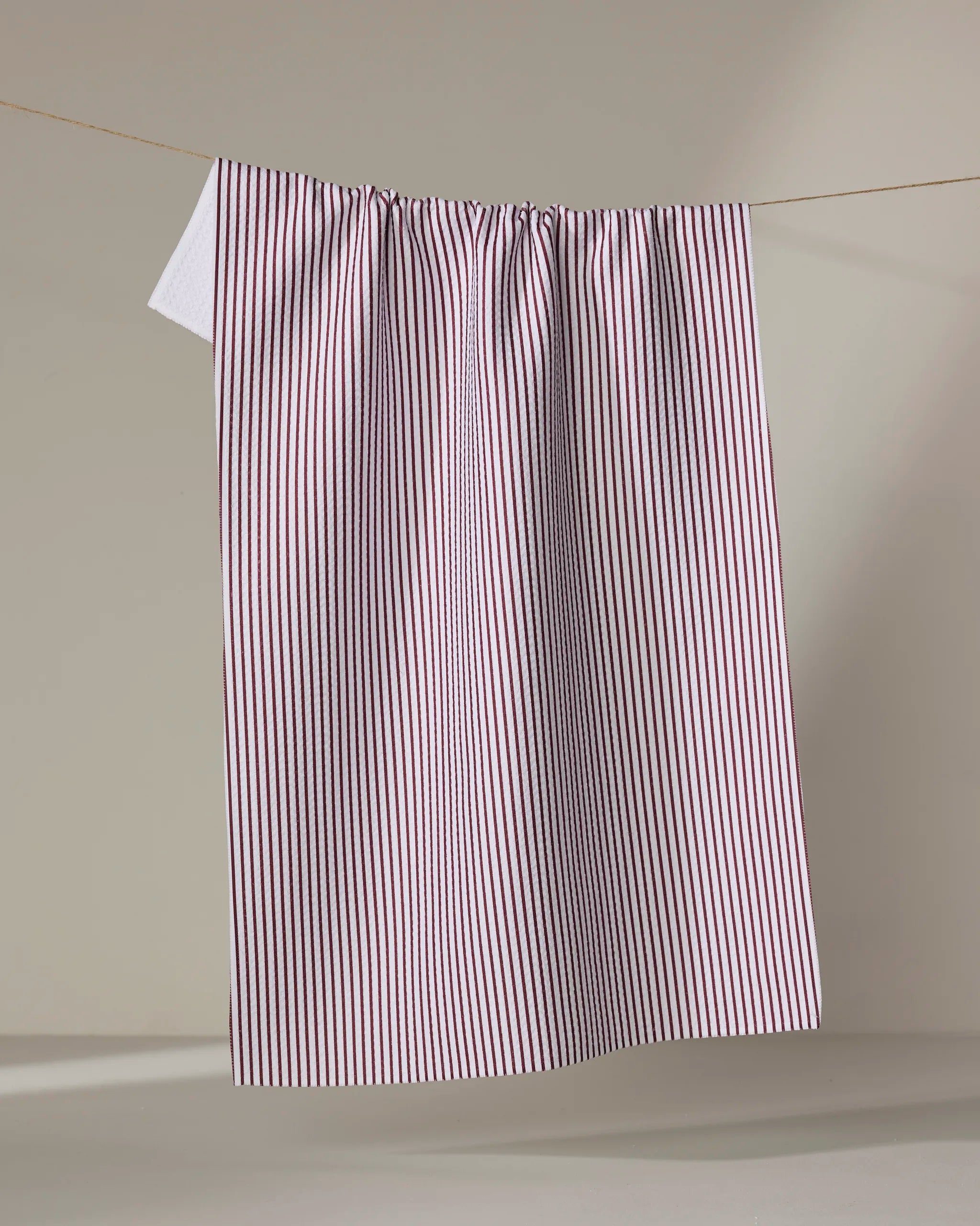 The Buy Guide's Get In Line - Wine tea towel, featuring red and white vertical stripes and ultra-absorbent fabric, hangs on a clothesline against a plain background, with a corner of another white cloth peeking behind it.