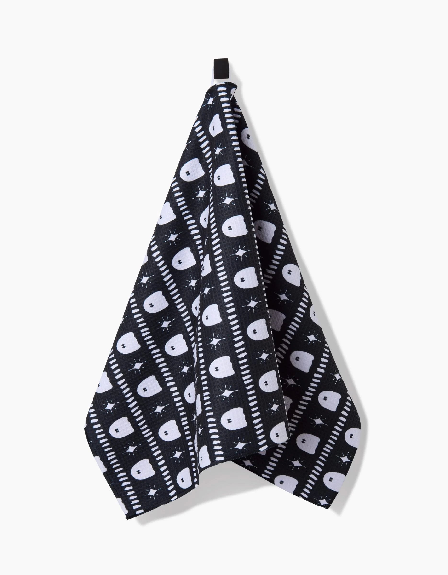 The Ghost Pac Tea Towel by Charlie Rowan Designs features white skulls, bones, stars, and diagonal stripes on black Geoweave™ fabric. It hangs by a black loop against a white background.