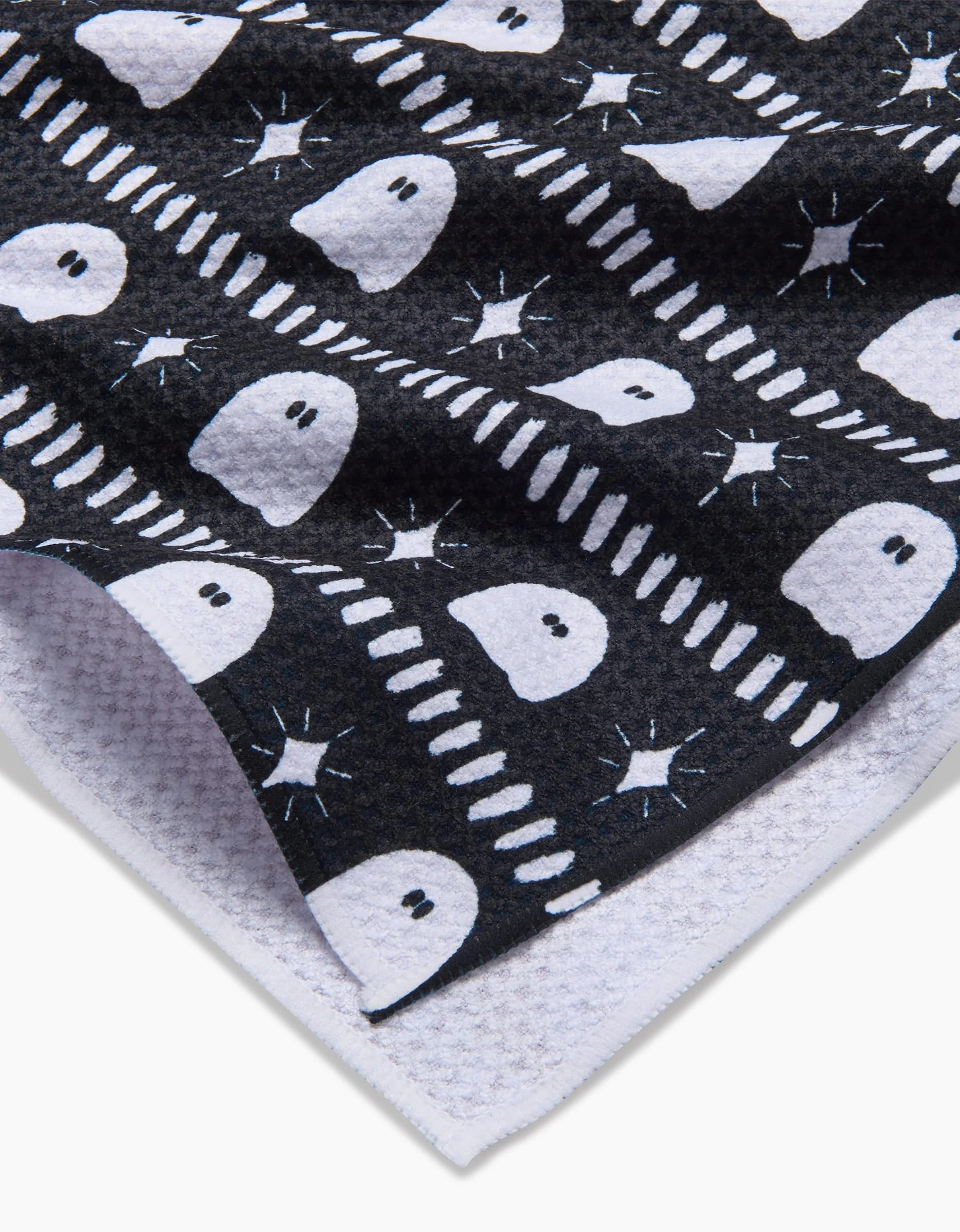 A close-up of the Ghost Pac tea towel by Charlie Rowan Designs, showing its black Geoweave™ fabric with a repeating pattern of white ghosts and stars, plus the textured edge revealing a white underside.