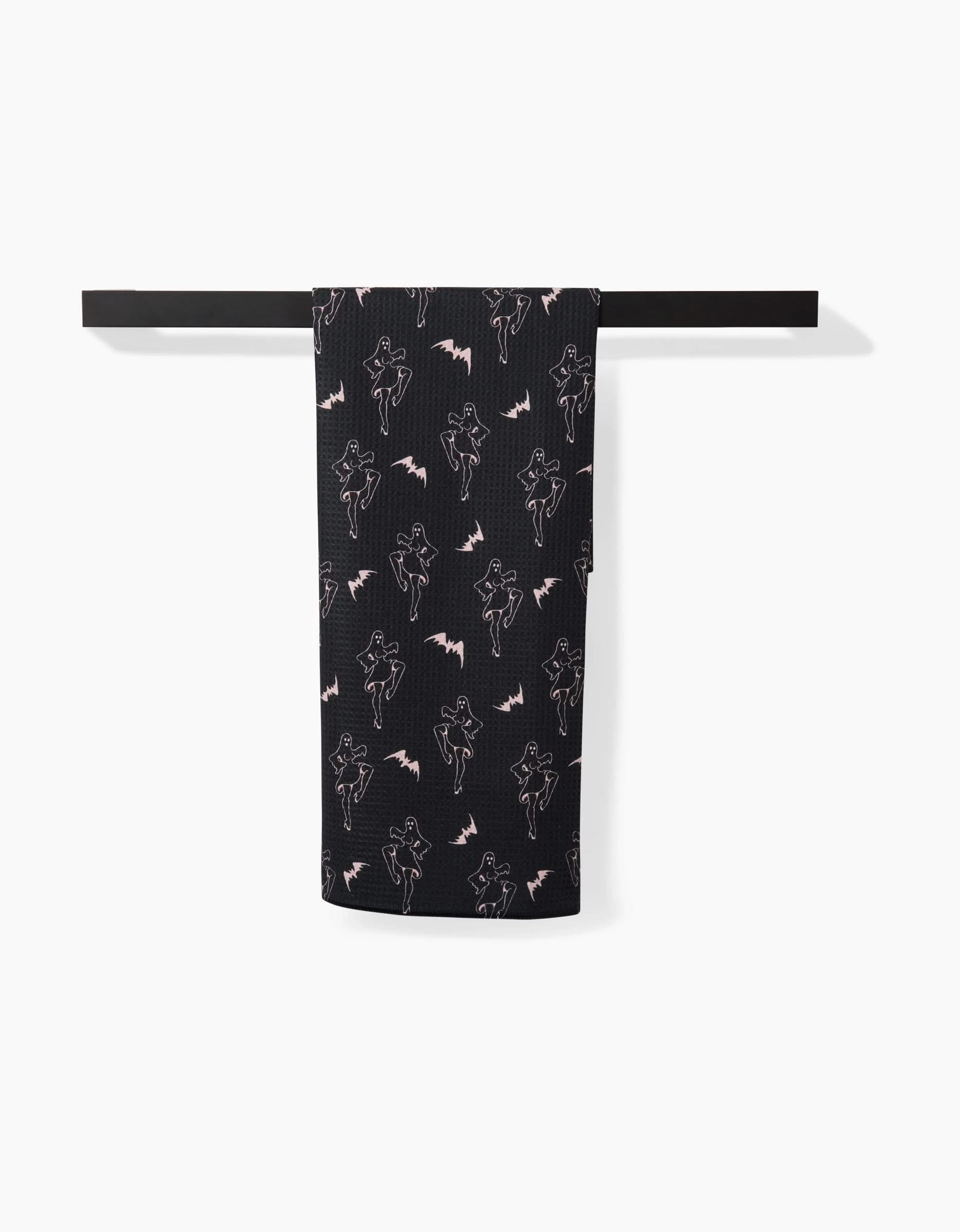 The Cole Strem Ghost Pin Up black tea towel hangs on a black rod, decorated with a repeating white outline of a dancing figure and small bats, and is crafted from recycled materials.