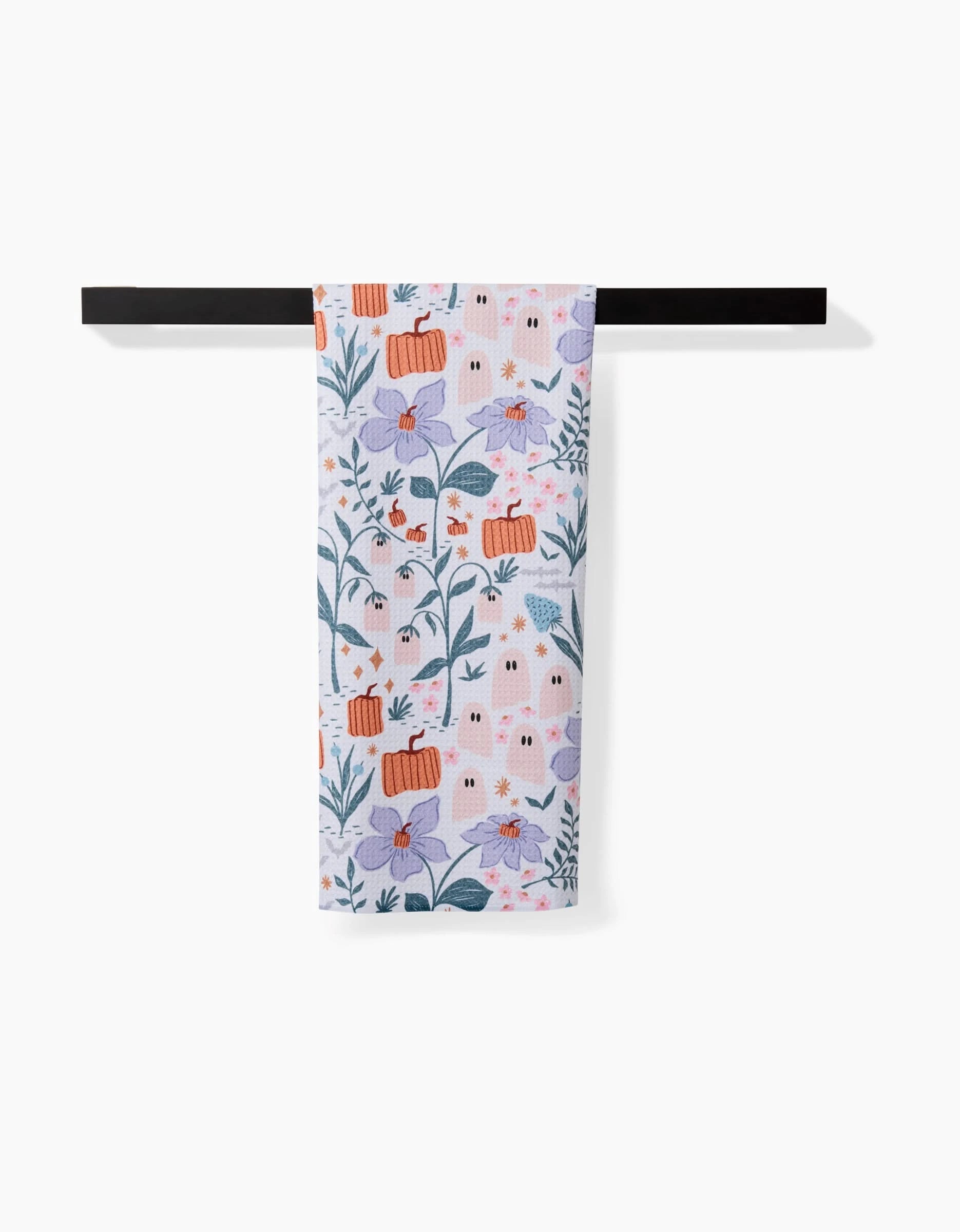 The Ghosts In The Garden tea towel by Charlie Rowan Designs, featuring orange pumpkins, purple flowers, green leaves, and ghost patterns, hangs on a black towel bar against a white background.