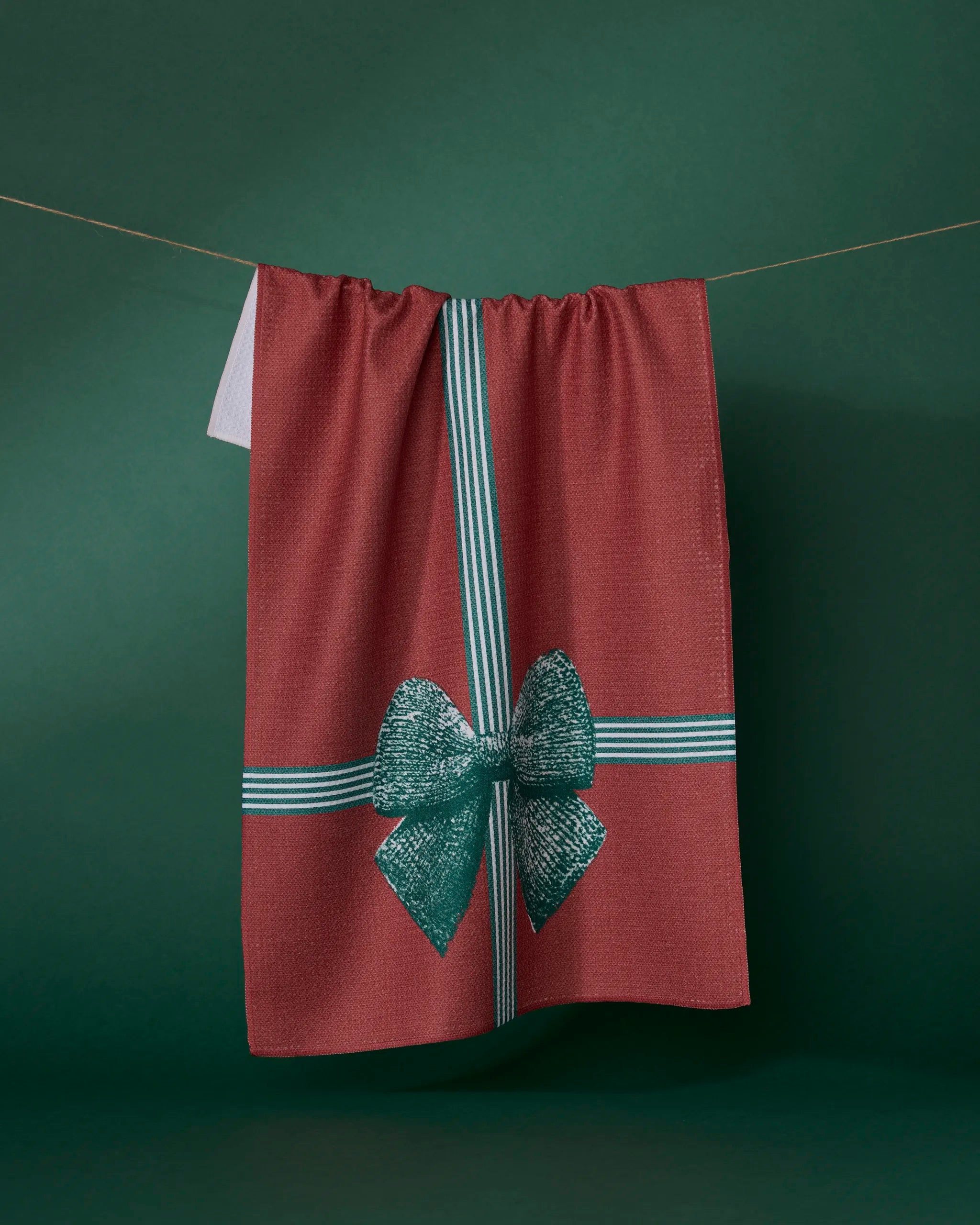 The Geometry "Gifted" tea towel, featuring a green ribbon and bow design on red, hangs against a green background and is made from recycled materials.