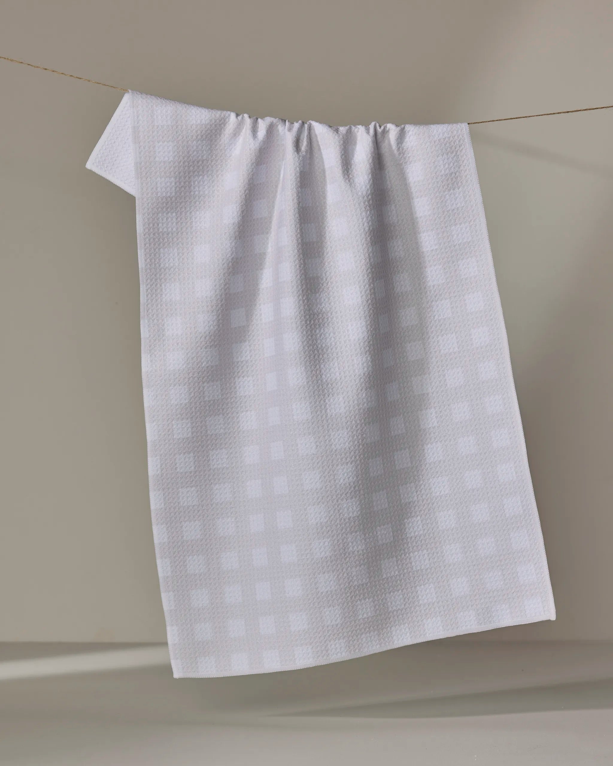 The Gigi - Beige kitchen towel by The Buy Guide hangs from a thin clothesline against a neutral backdrop, casting a soft shadow on the wall.