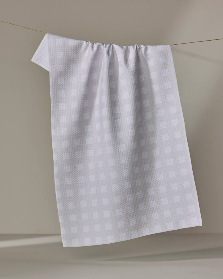 The Gigi - Beige kitchen towel by The Buy Guide hangs from a thin clothesline against a neutral backdrop, casting a soft shadow on the wall.