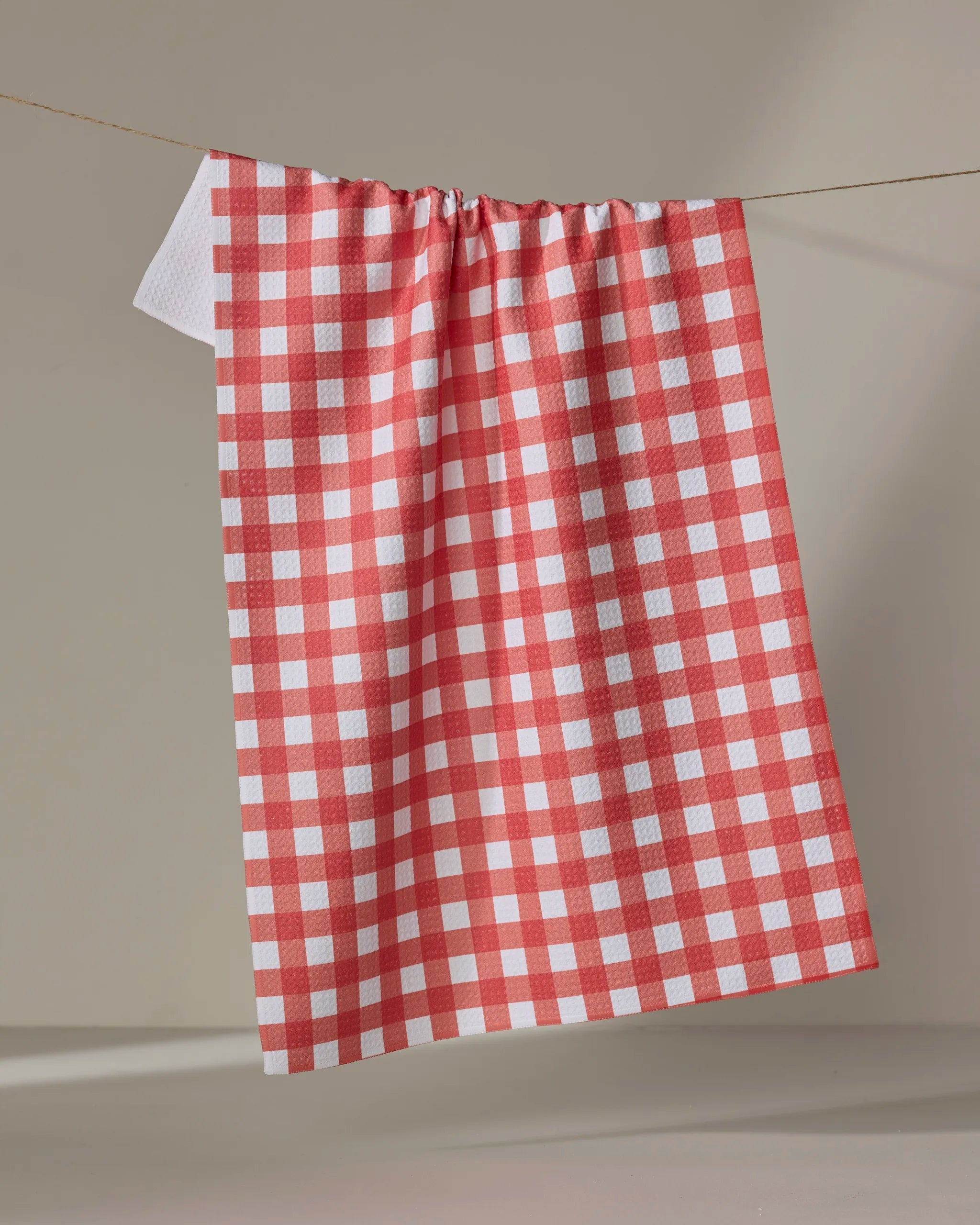 The Gigi - Candy Apple kitchen towel by The Buy Guide, featuring red and white checks, hangs draped over a clothesline against a neutral backdrop, displaying soft folds and eco-friendly style.