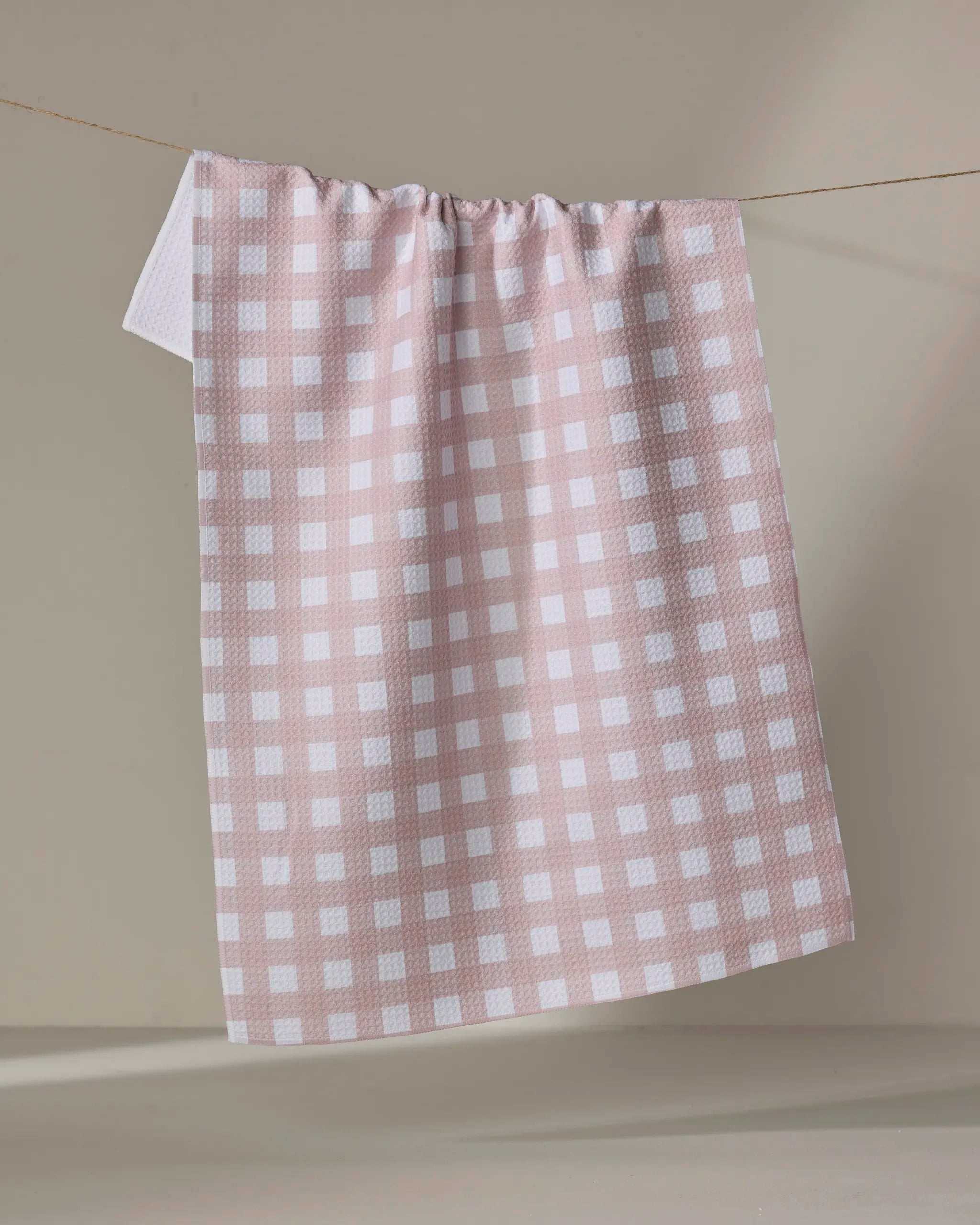 The Buy Guide's Gigi - Dusty Rose tea towel hangs on a string against a neutral beige background, bringing charm and practical style to any kitchen.