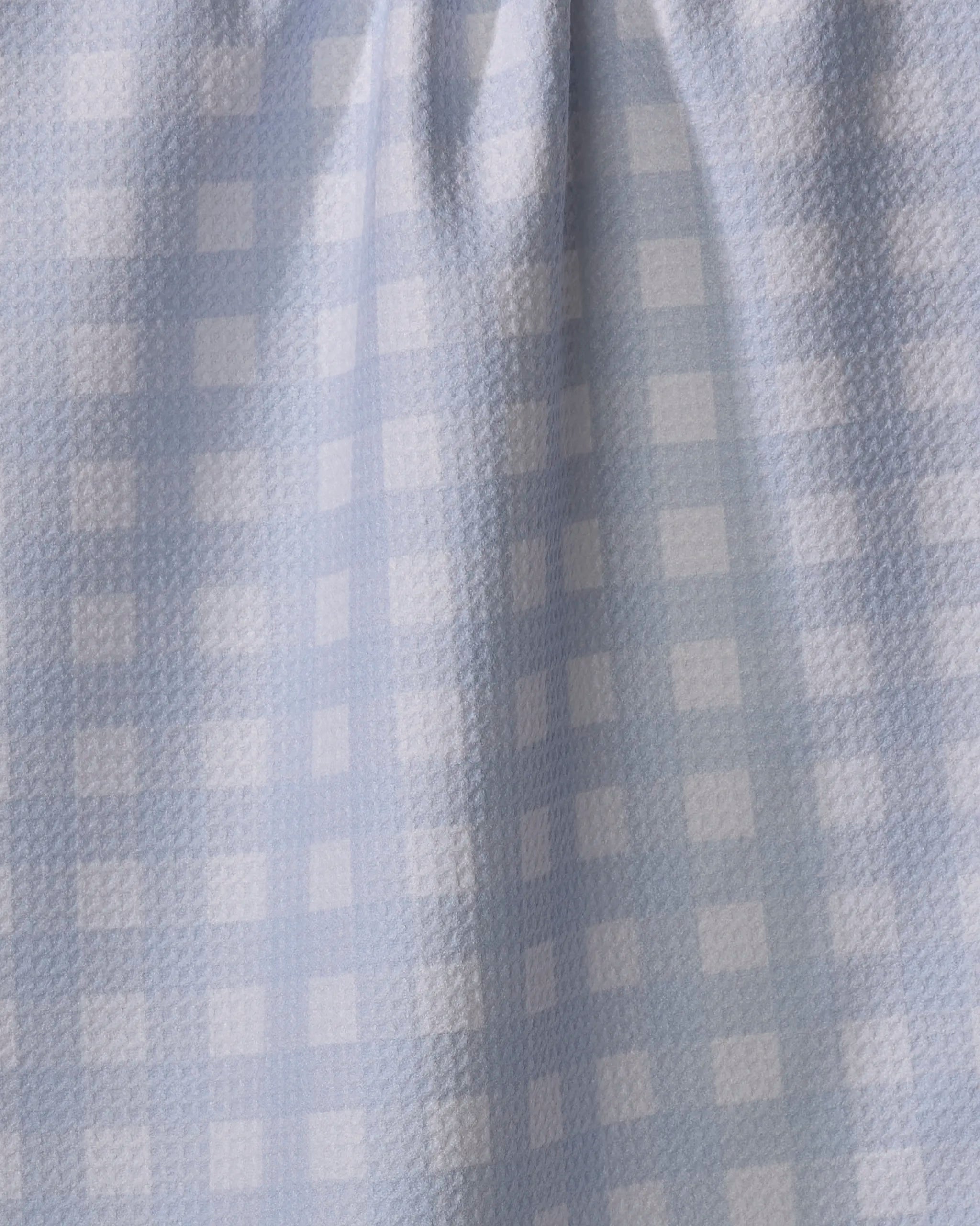 The Gigi - Light Blue dish towel by The Buy Guide features light blue and white checkered fabric with a subtle texture, soft folds, and absorbent material that creates gentle shadows and highlights across the surface.