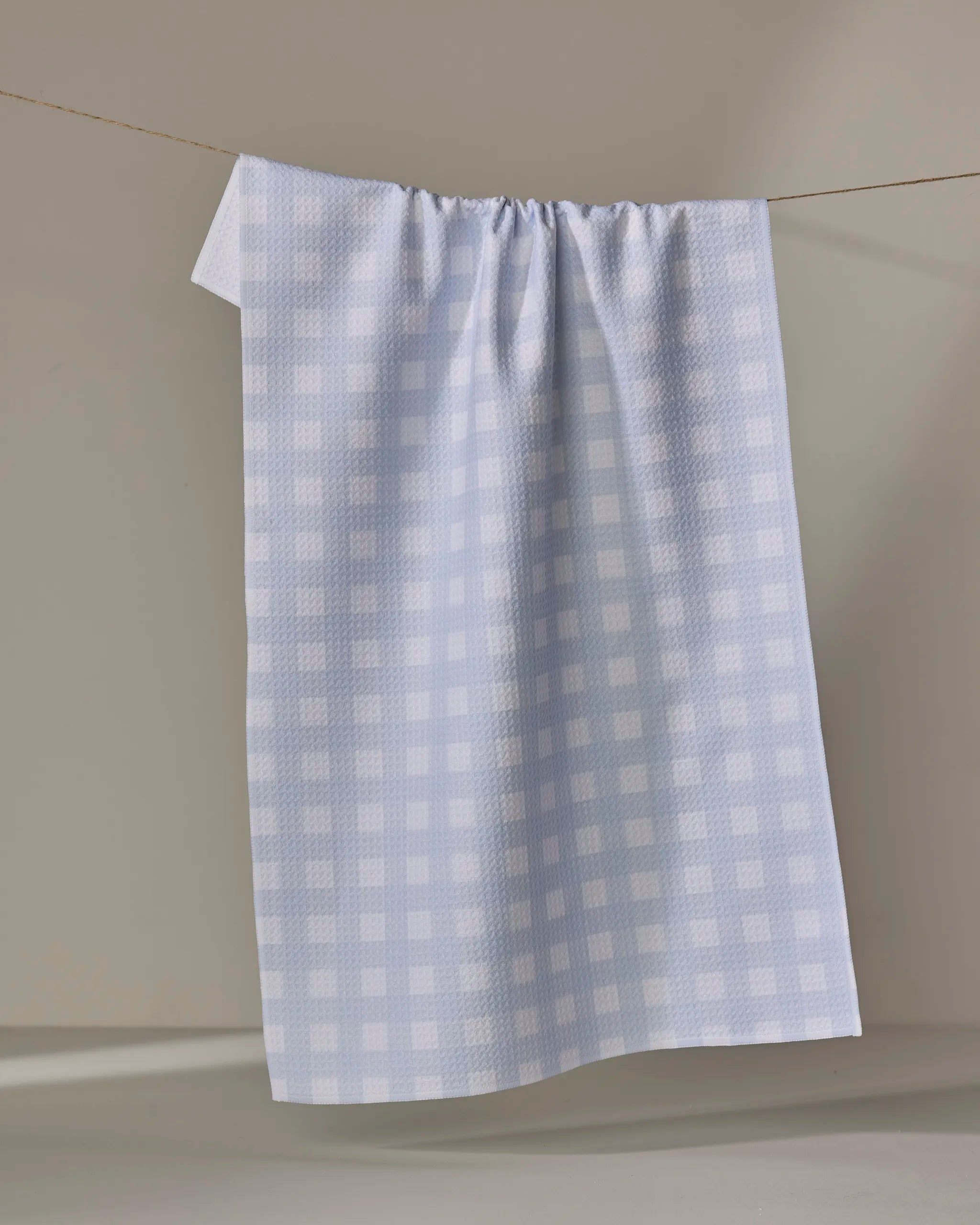 The Buy Guide’s Gigi - Light Blue tea towel hangs on a thin clothesline, displaying its light blue and white checkered pattern against a neutral background.