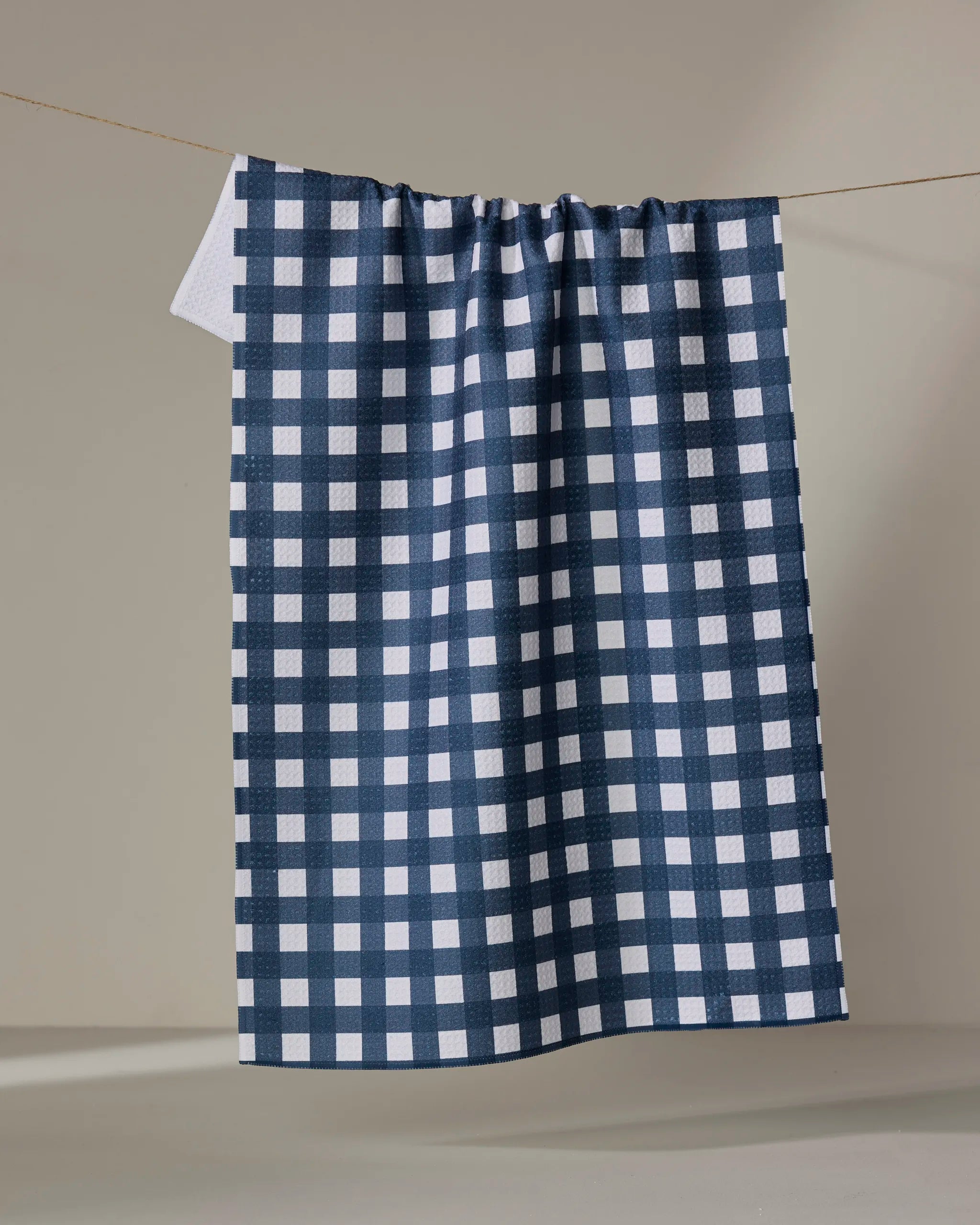 The Gigi - Navy tea towel by The Buy Guide, featuring a navy blue and white checkered pattern, hangs neatly on a clothesline against a plain beige background.