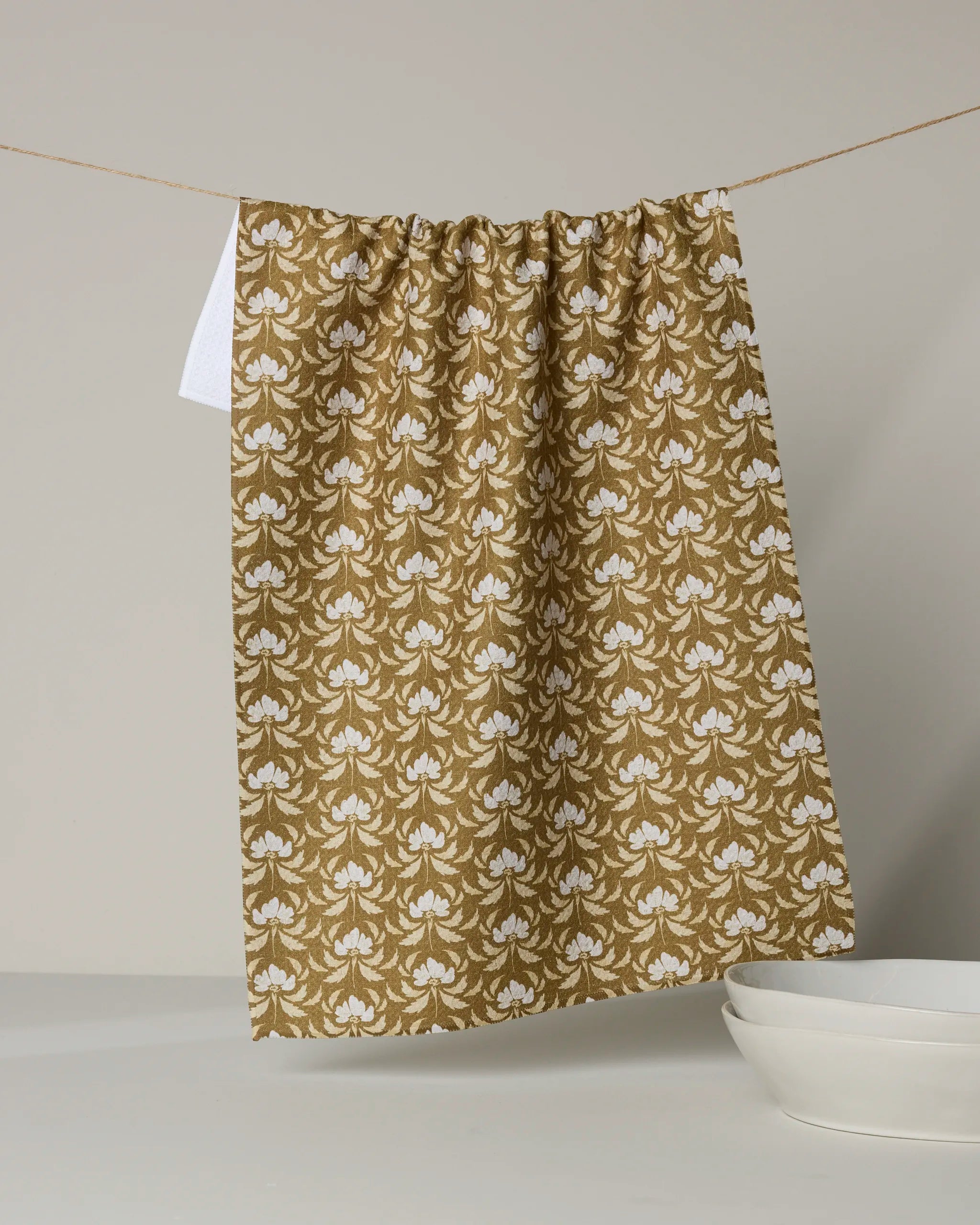 The Gilded Bloom recycled kitchen towel by Melissa Johnson, featuring a gold and white pattern, hangs on a clothesline above a white counter with two stacked white bowls below.