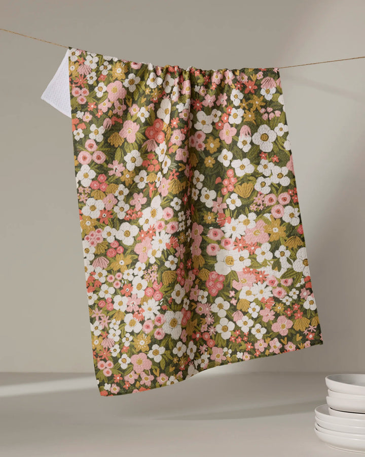 The “Gilded Garden” tea towel by Lisa Nelson, featuring a pink, white, and green floral print, hangs on a line while white plates are stacked in the corner against a neutral backdrop.