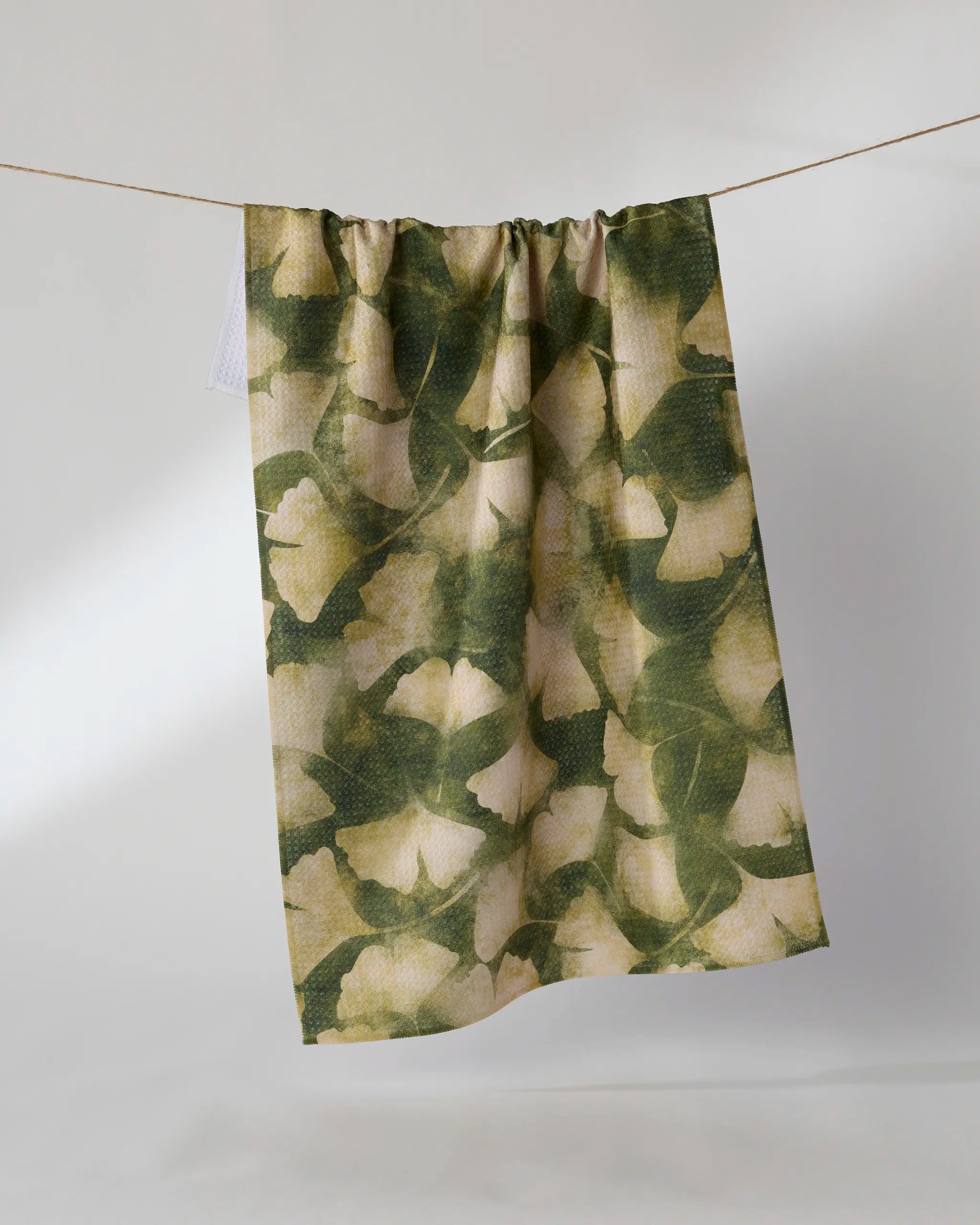 The Geometry Gilded Ginkgo tea towel, featuring green and beige floral patterns, is displayed hanging on a string against a plain white background.