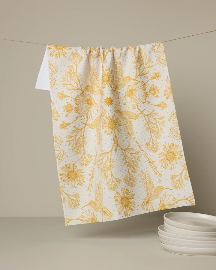 The Geometry Gilded Nectar tea towel, featuring a yellow floral and hummingbird design, hangs above a beige surface next to a stack of white plates.