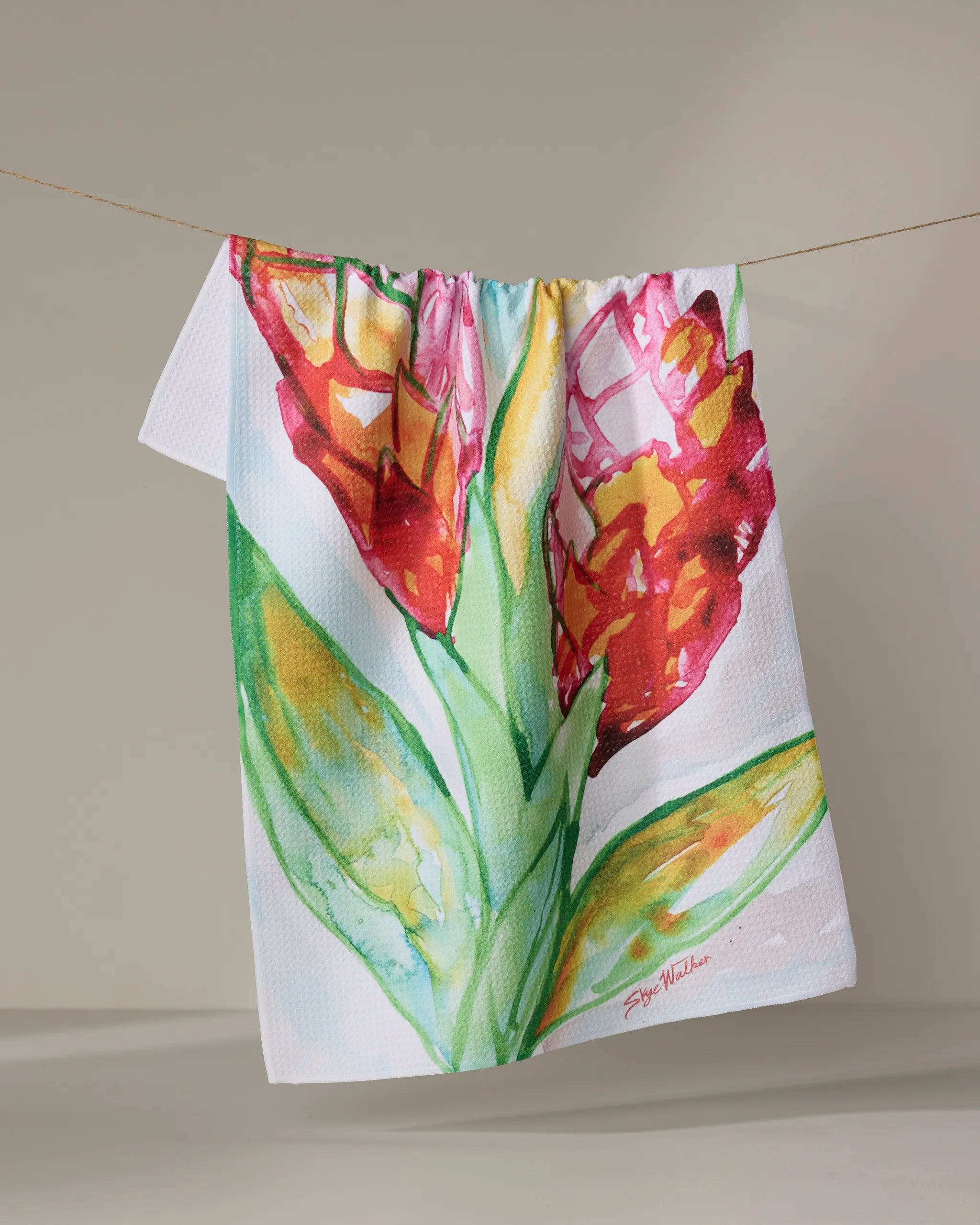 The Skye Walker "Ginger" kitchen towel is an eco-friendly white towel featuring a vibrant watercolor red and yellow flower with green leaves, shown hanging on a clothesline against a neutral background.