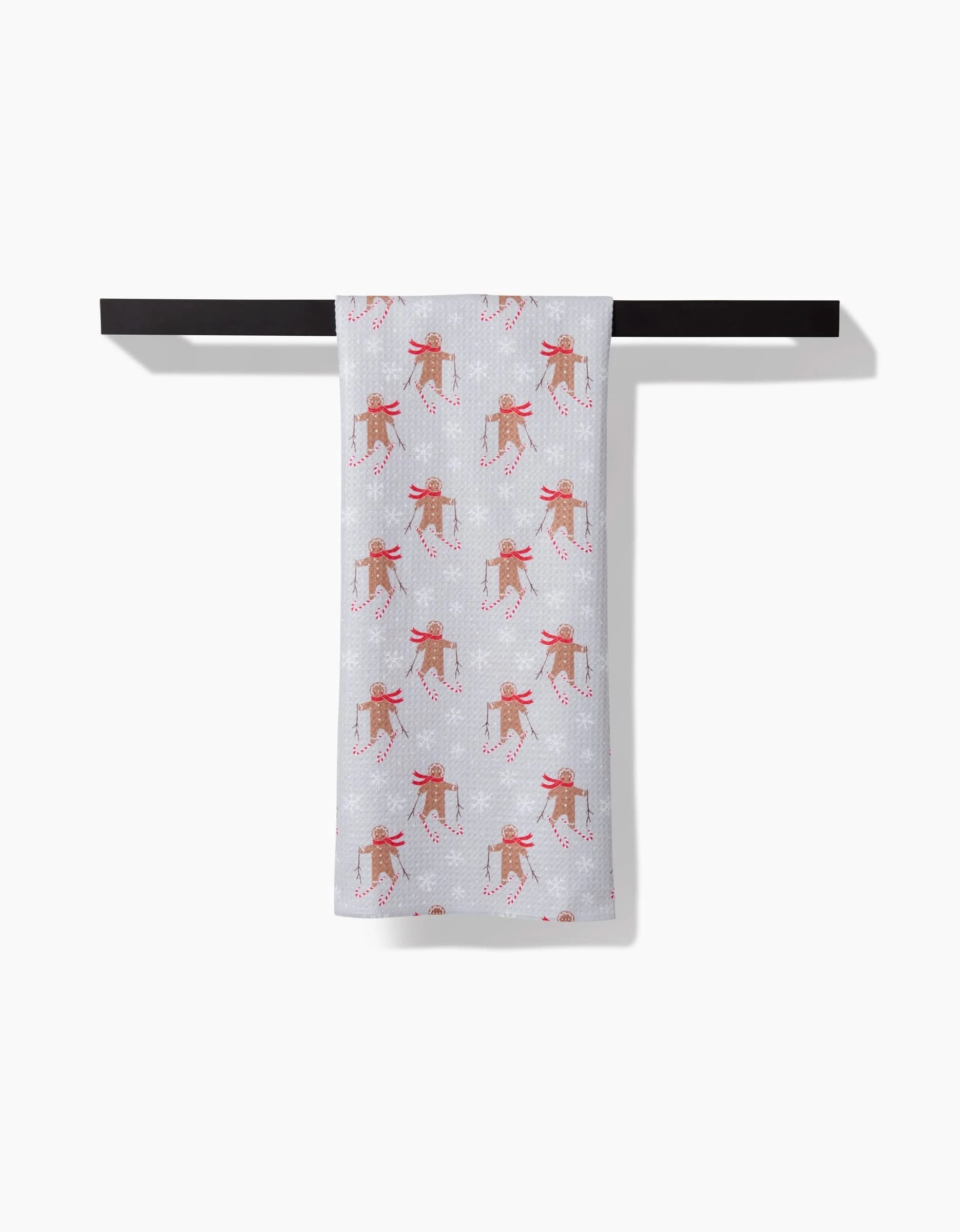 The Roxy Marj Gingerbread Skiers tea towel features ultra-absorbent white fabric with skiing reindeer in red scarves, displayed neatly over a black towel bar against a white backdrop.