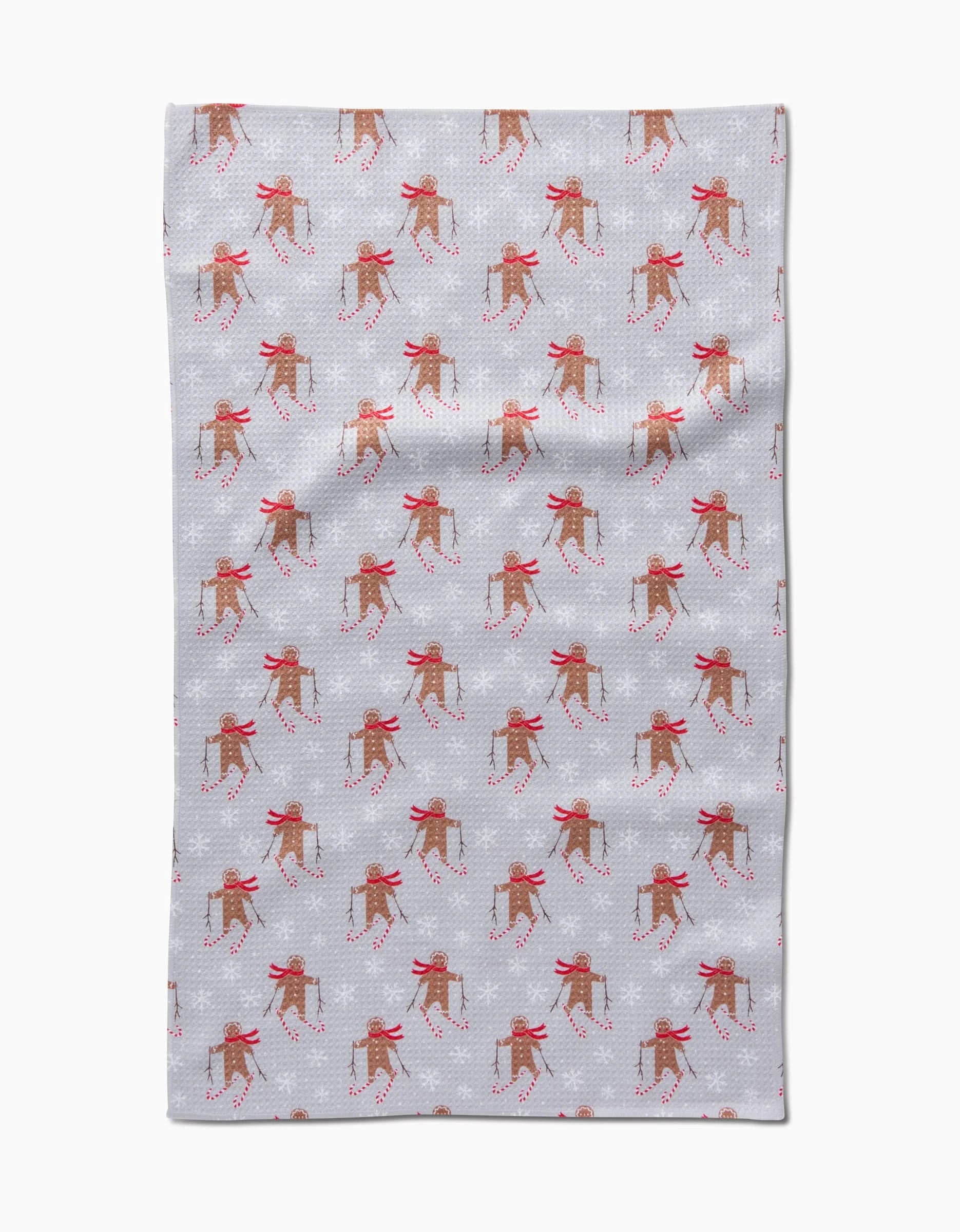 The Roxy Marj Gingerbread Skiers tea towel is light gray and decorated with a repeating pattern of reindeer in red scarves holding candy canes, set against a subtle dotted background.