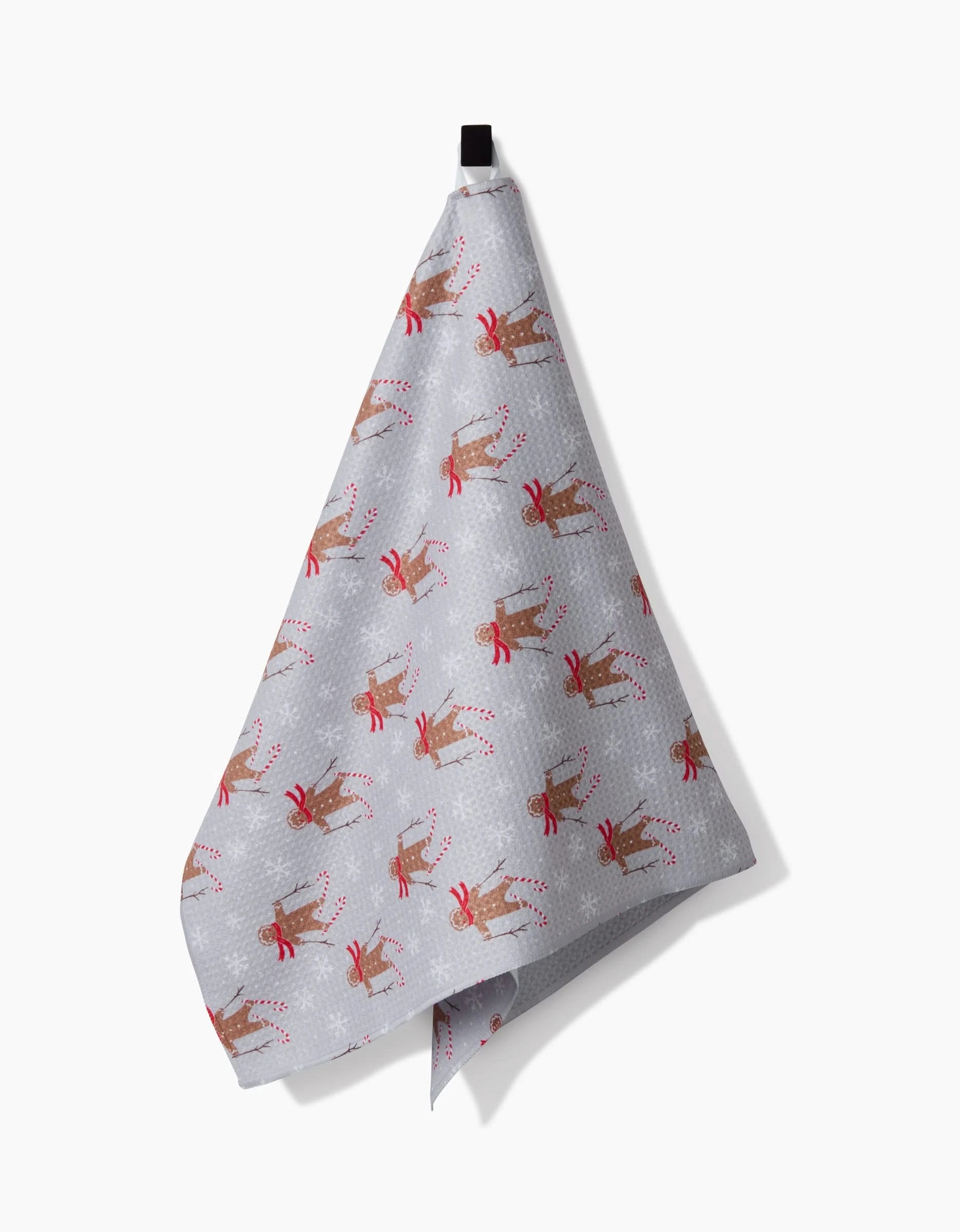 A light gray kitchen tea towel by Roxy Marj, called Gingerbread Skiers, featuring gingerbread skiers in red scarves and snowflakes, hanging from a black loop against a white background.
