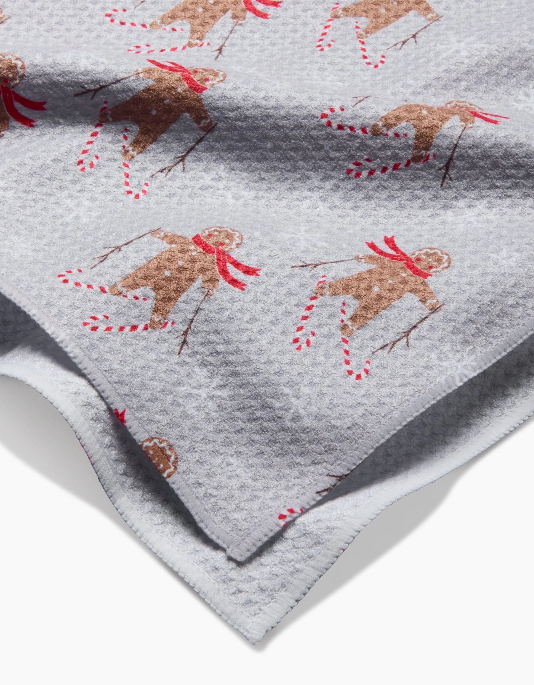 The Roxy Marj Gingerbread Skiers tea towel is light gray with a textured finish, featuring gingerbread people skiing in red scarves on candy cane skis.