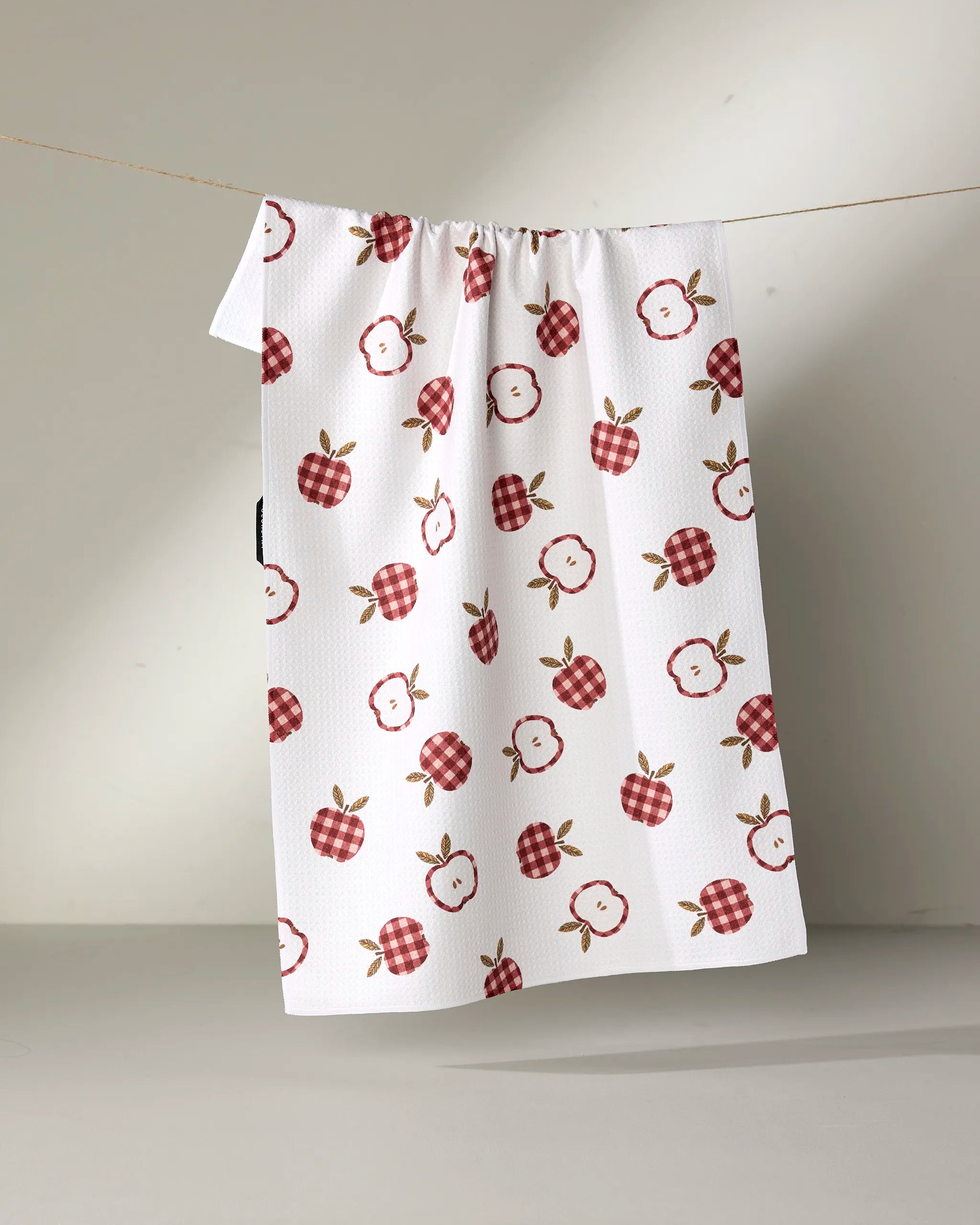 The "Gingham Apples" kitchen towel by Teri the Butter showcases red and plaid apples with slices, brown stems, and green leaves on a light background.