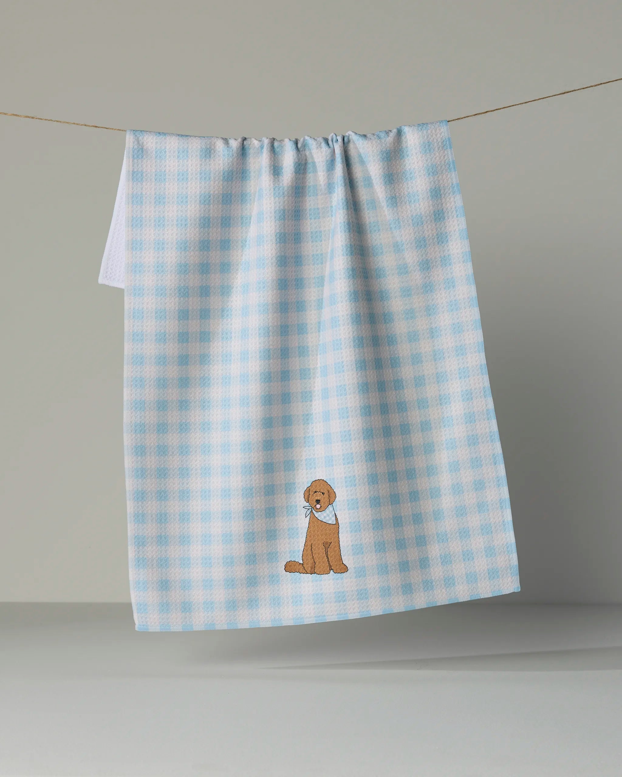 A blue and white checkered tea towel from Geometry, the Gingham Doodle, hangs on a line. Made from recycled materials, it features an embroidered brown dog sitting near the bottom center.