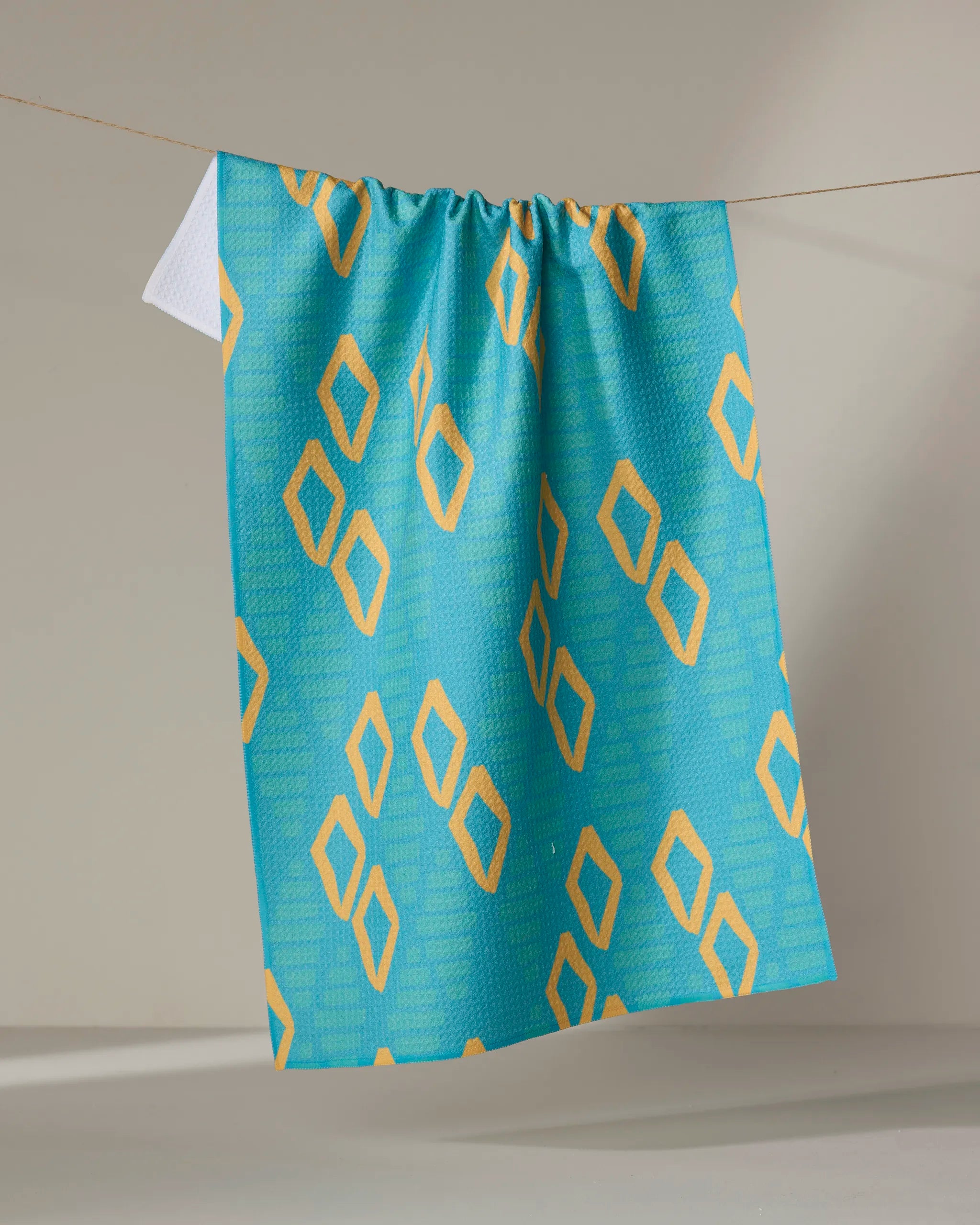 The "Girl's Best Friend" tea towel by Sareka Unique features a blue and yellow diamond pattern, hanging stylishly from a string against a neutral backdrop to brighten any kitchen space.