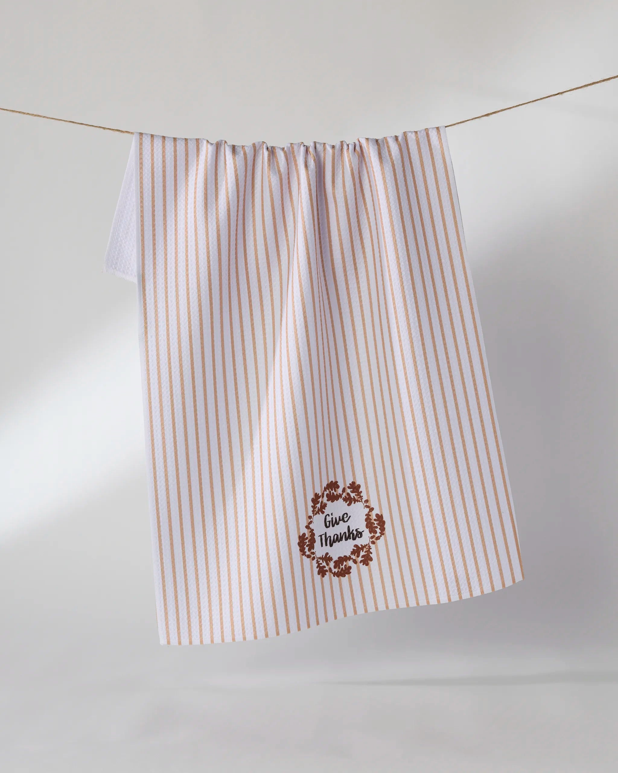 The Geometry "Give Thanks" tea towel features vertical tan stripes on white, a small wreath with "Give Thanks" printed in the corner, and is made from recycled materials. It hangs neatly on a line.