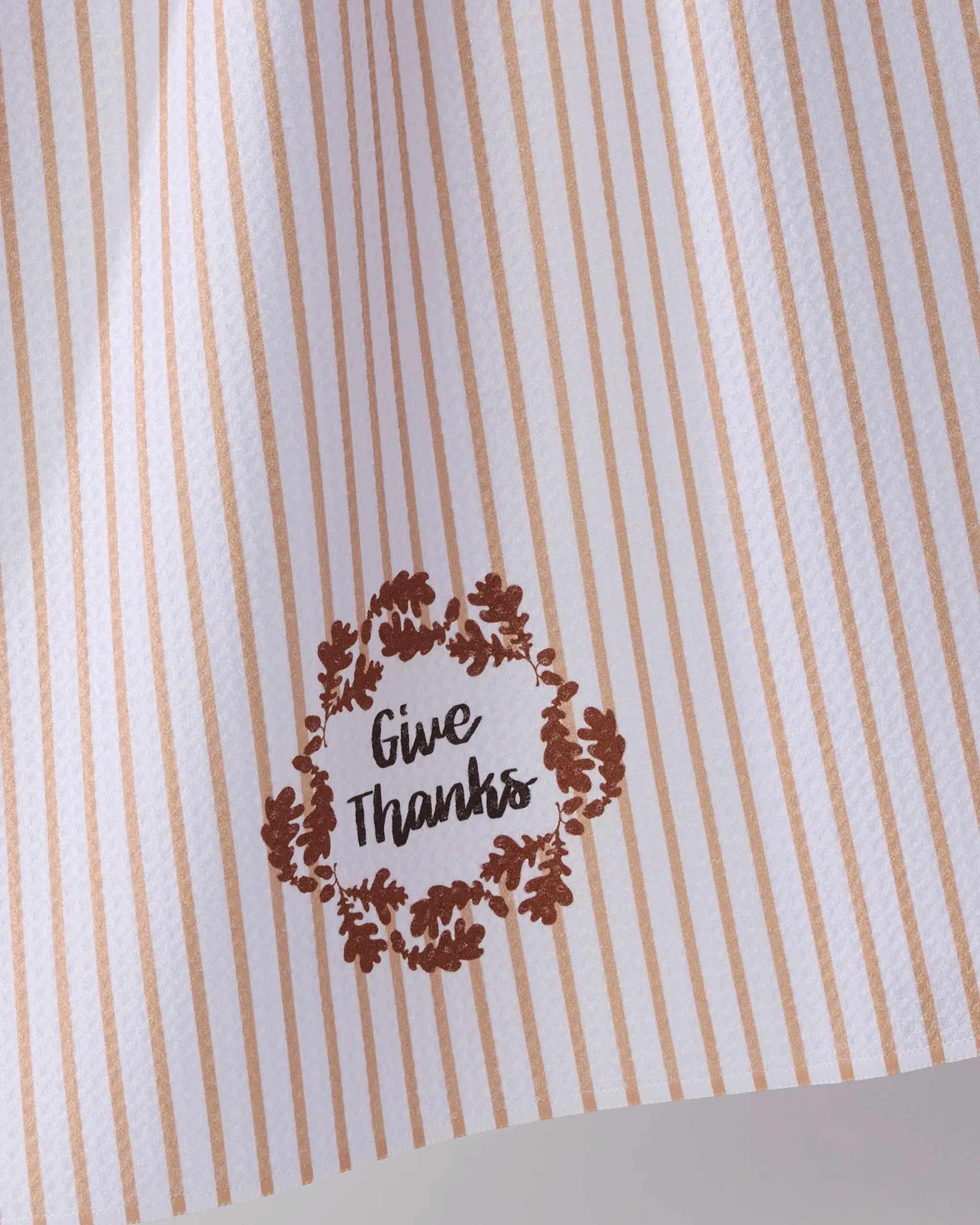 The Geometry "Give Thanks" tea towel features light brown vertical stripes and a brown leafy wreath with "Give Thanks" in the corner. Made from recycled materials, it offers an eco-friendly touch for your kitchen.
