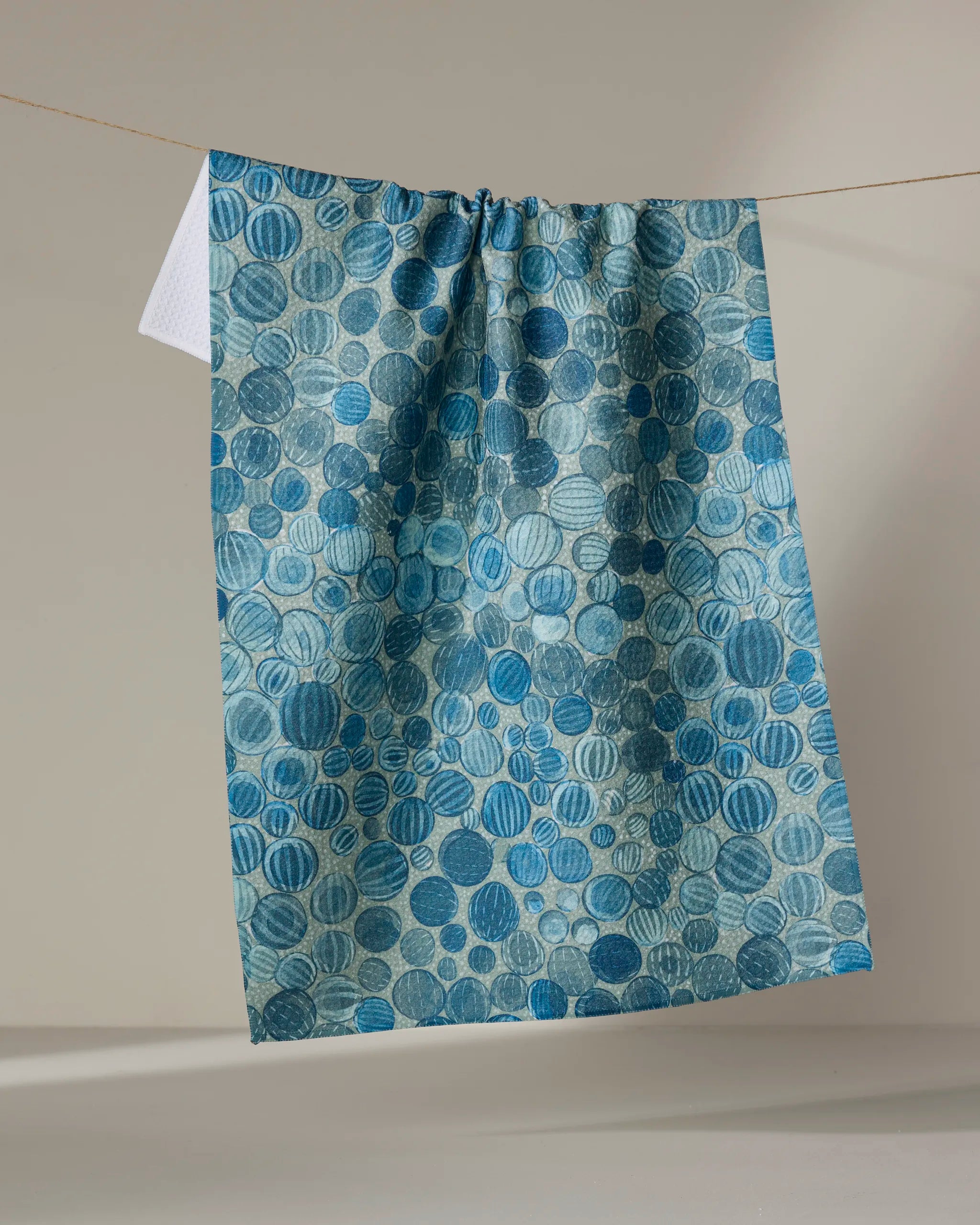 A blue ultra-absorbent tea towel, Glass Baubles by Katie O'Shea, featuring variously shaded blue circles, hangs on a clothesline against a neutral background.