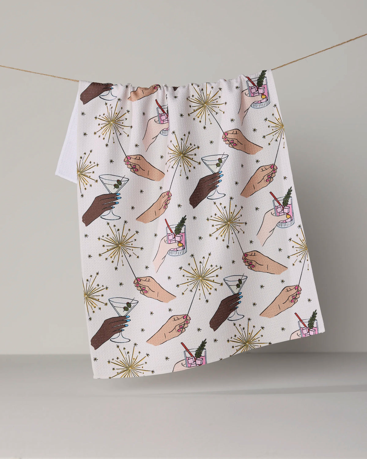 Here’s a revised version of the sentence using the provided product data: The Geometry Glitter Cheers recycled kitchen towel hangs on a string, featuring illustrated hands holding cocktails and champagne glasses, accented with gold sparkler and star designs on a crisp white background.