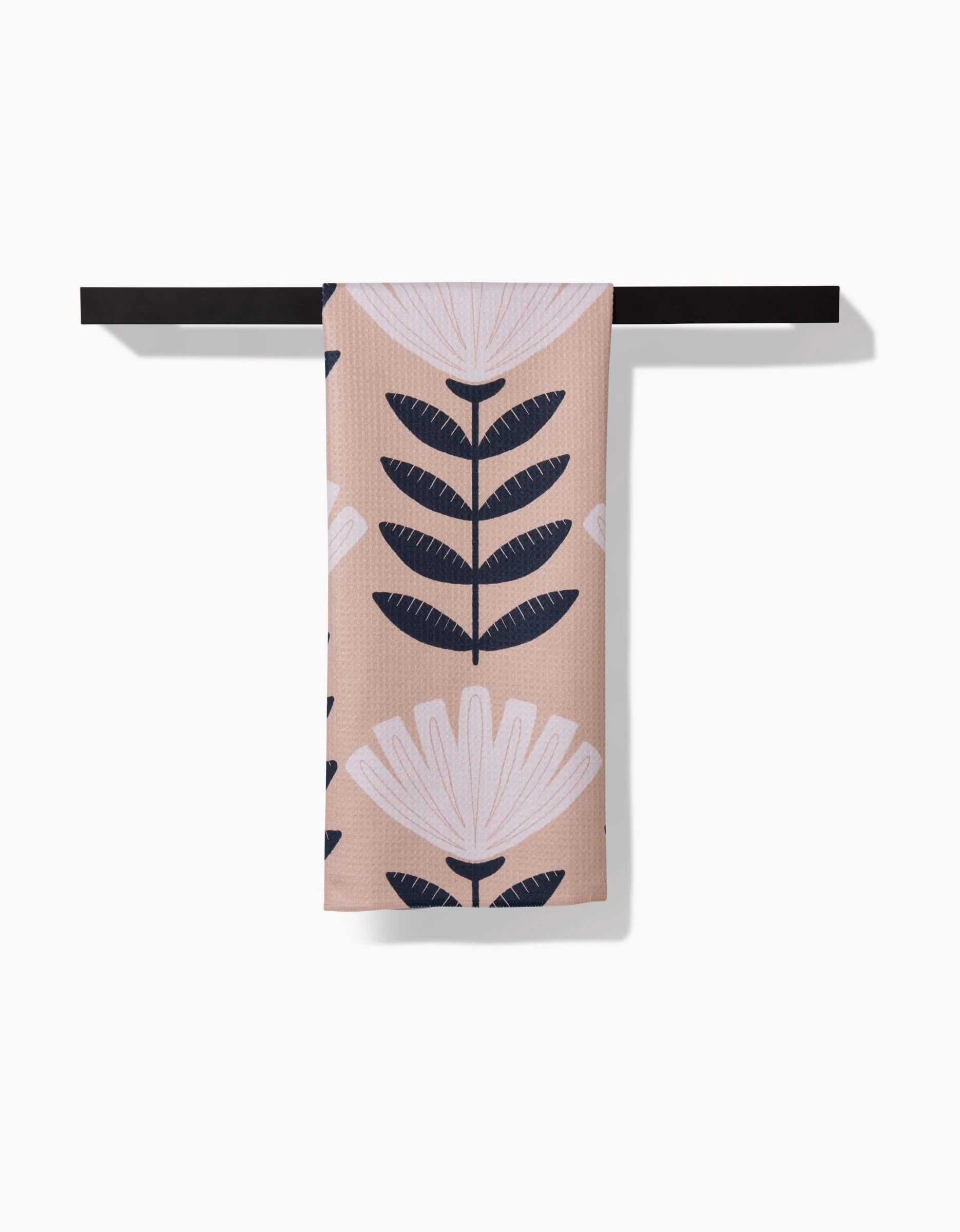 The Gloria towel by Amy MacCready, made from post-consumer recycled materials and featuring a white and dark blue floral design on beige, hangs neatly on a black towel bar against a white background.