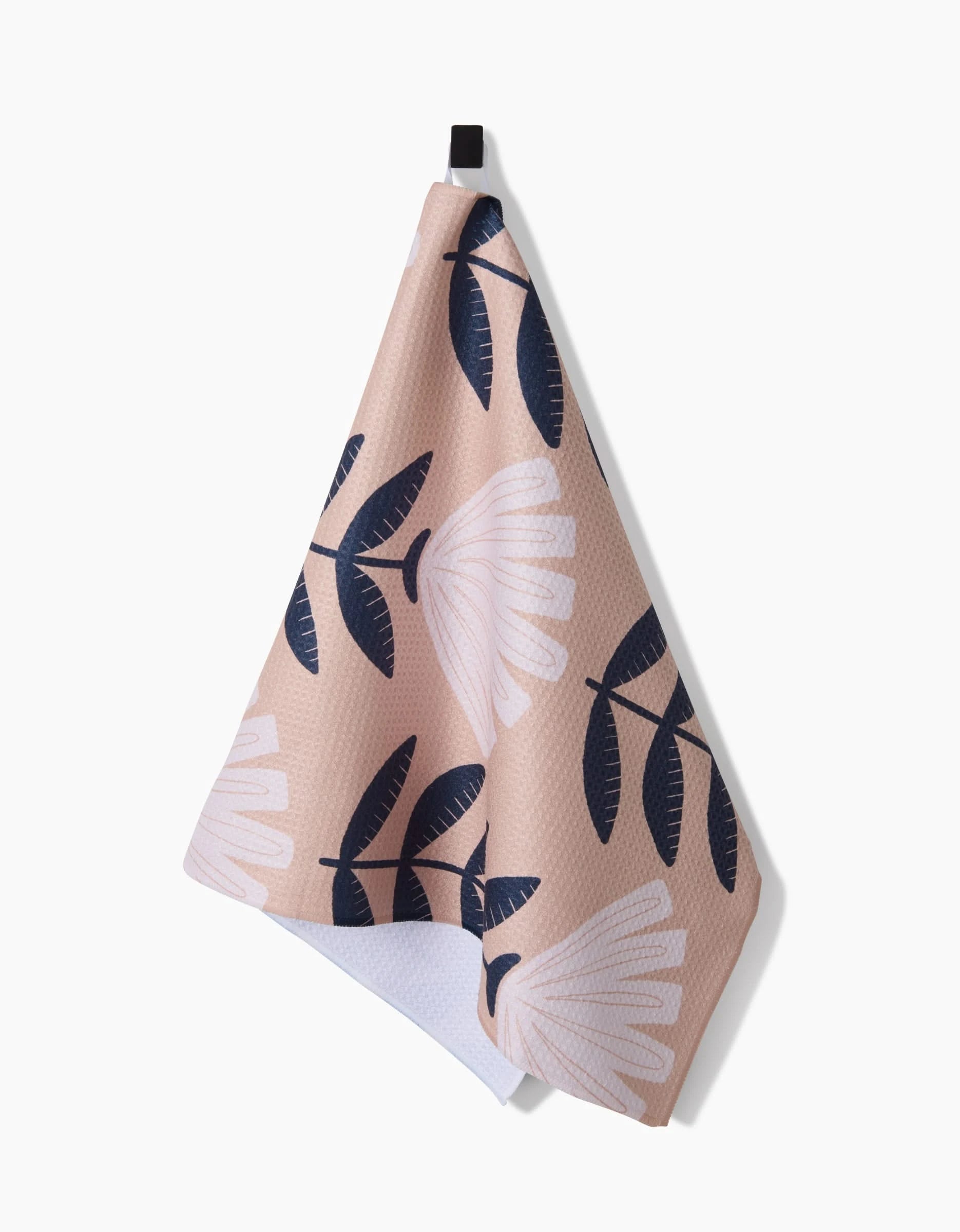 A peach-colored Gloria tea towel by Amy MacCready, made from Geoweave fabric with white flower and dark blue leaf patterns, hangs from a black loop against a white background.