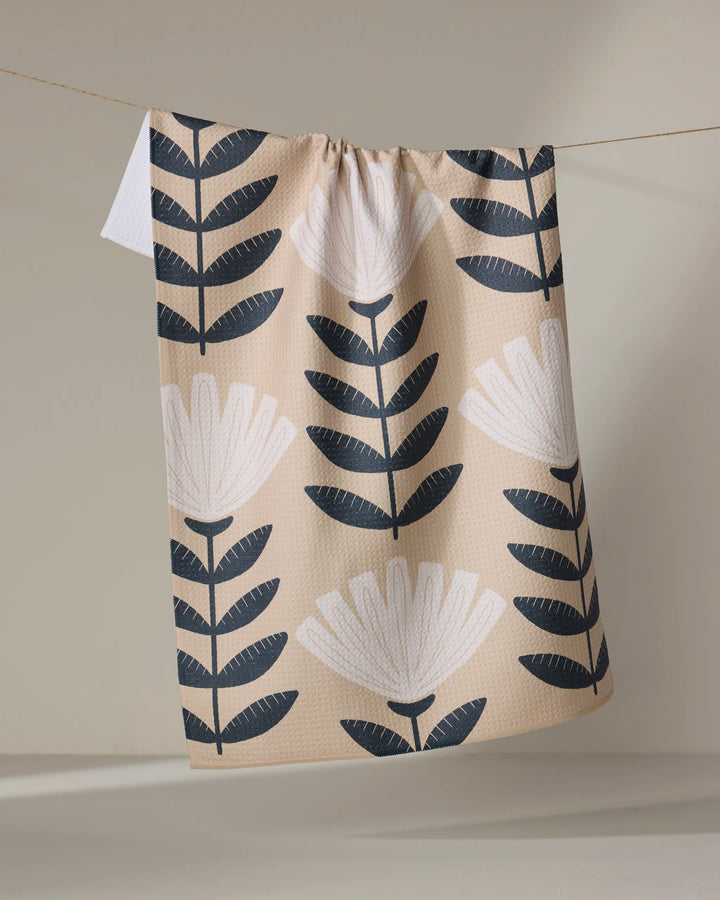 The Gloria kitchen tea towel by Amy MacCready, crafted from recycled materials, displays large black and white stylized flowers and leaves on beige fabric, hanging on a clothesline against a neutral backdrop.