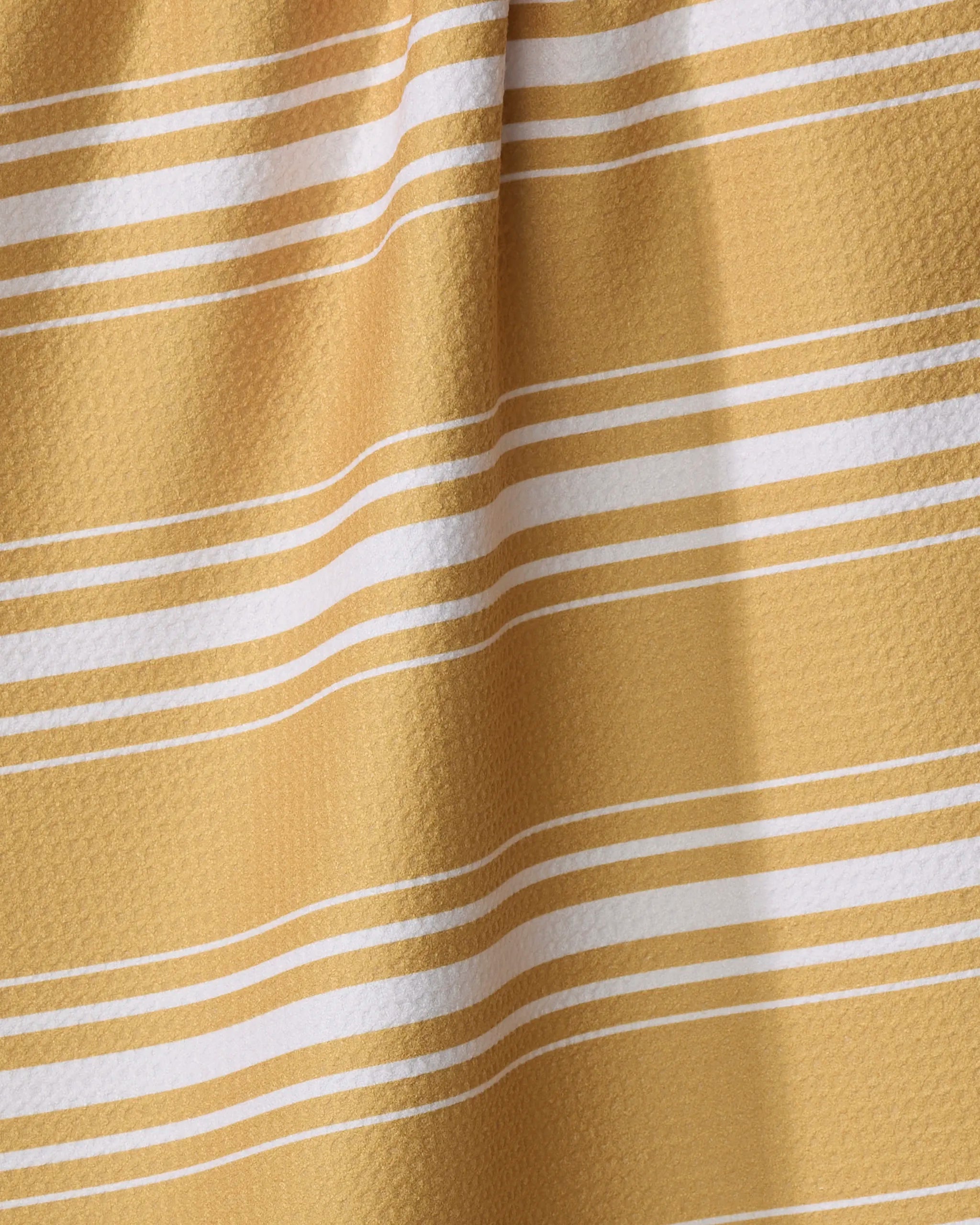 A close-up of the Katie O'Shea "Gold" tea towel, featuring yellow and white horizontal stripes on a textured surface with soft folds. Made from quick-drying, recycled materials.