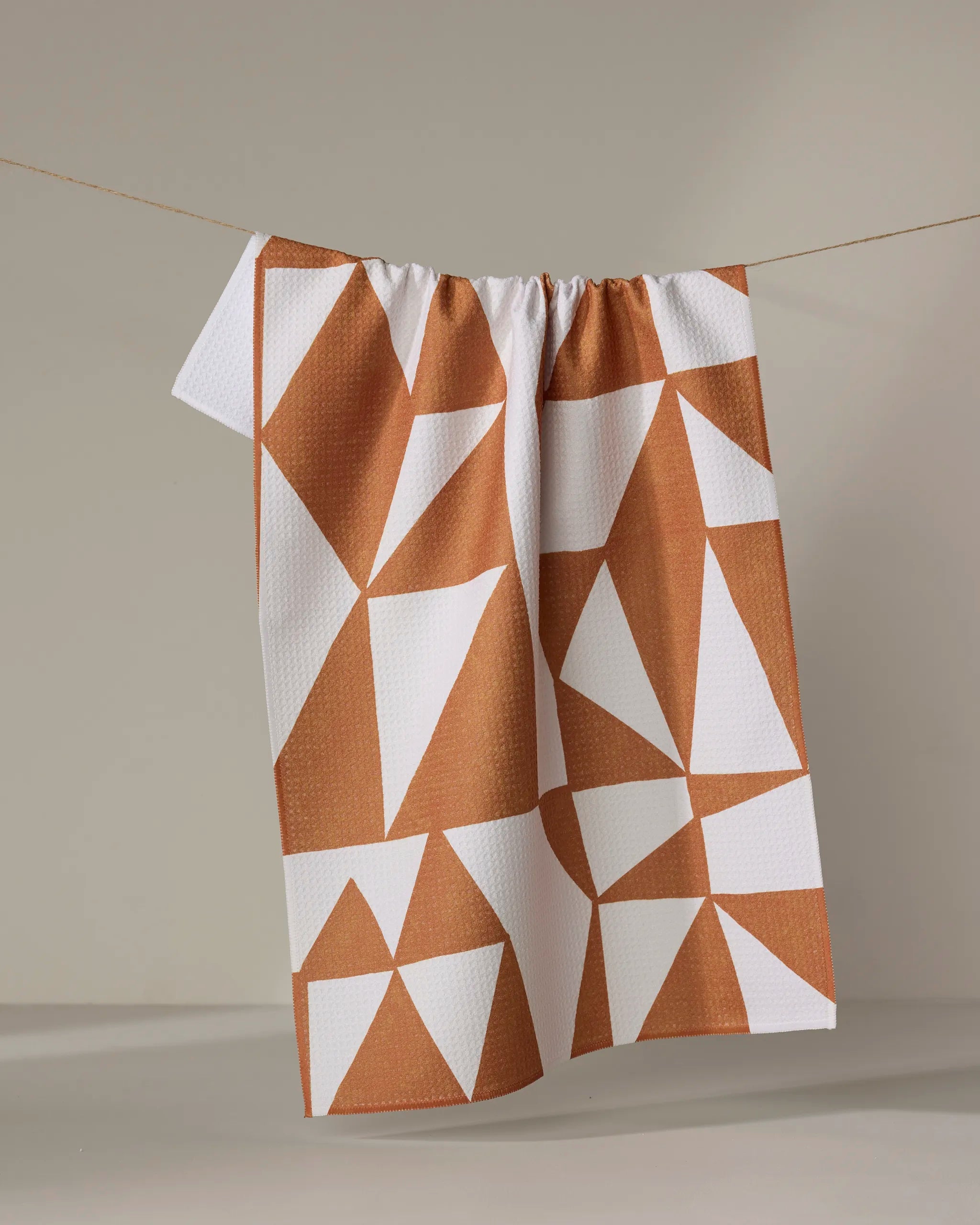 The "Golden Angles" tea towel by Lisa Nelson, featuring a geometric white and terracotta triangle pattern and crafted from recycled materials, hangs on a clothesline against a neutral background.