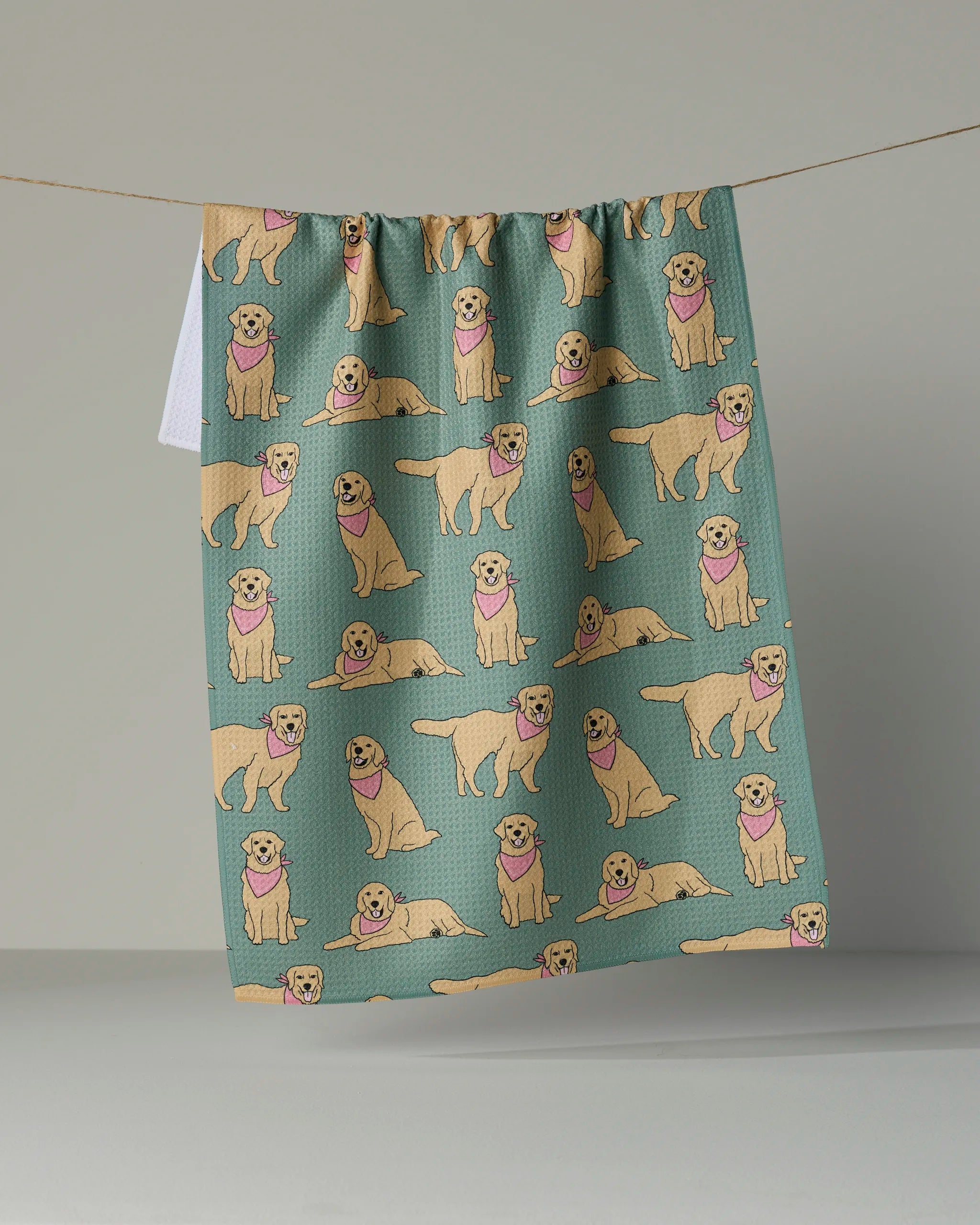 A Geometry “Golden Days” ultra-absorbent green towel, featuring a repeating pattern of golden retriever illustrations—some sitting, some lying down, and some wearing pink bandanas—hangs on a clothesline against a plain background.
