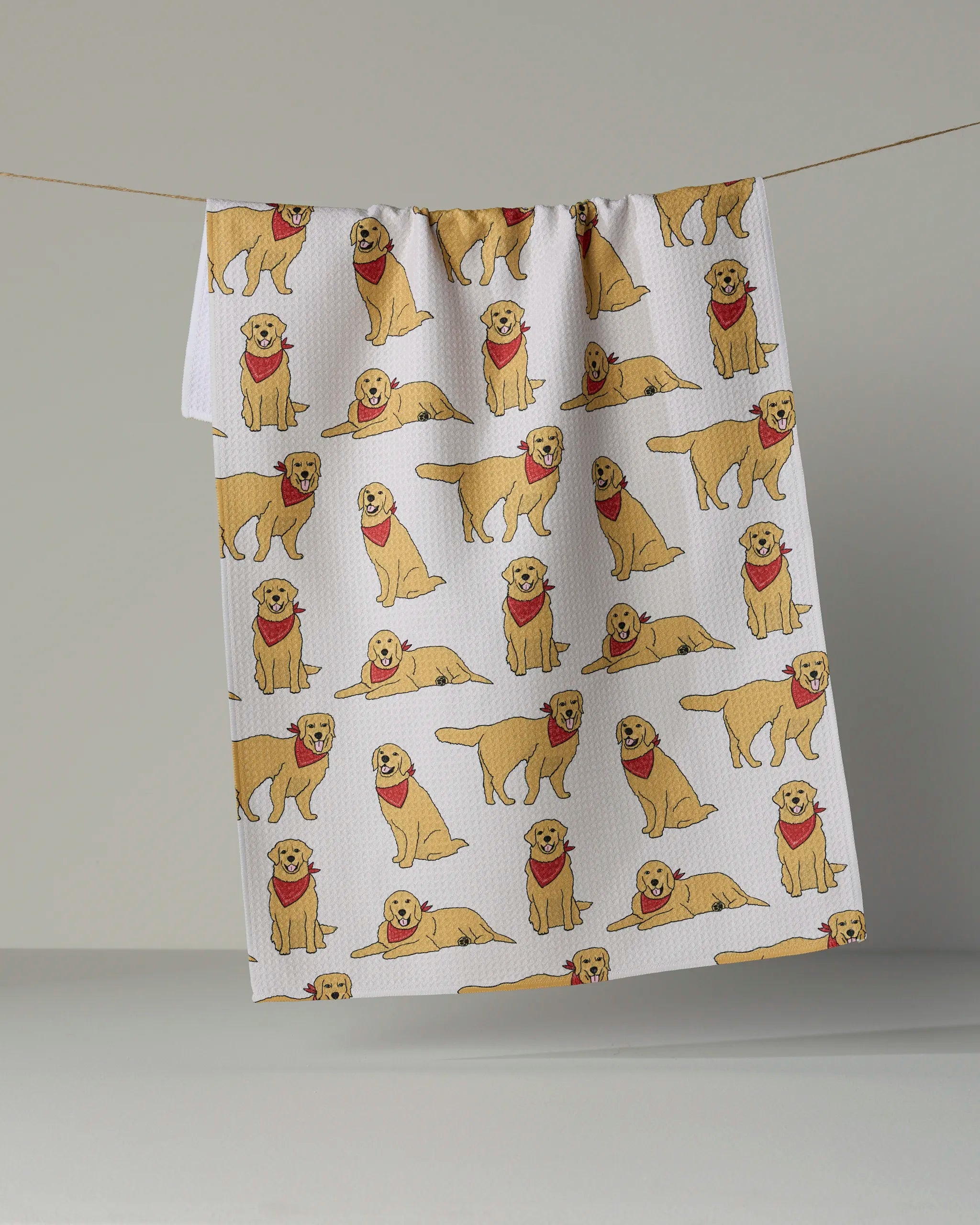 The Geometry “Golden Days” tea towel, crafted from recycled materials, displays a repeating pattern of yellow dogs in red bandanas. Shown hanging on a line against a neutral background, this white kitchen towel combines playful design with eco-friendly style.