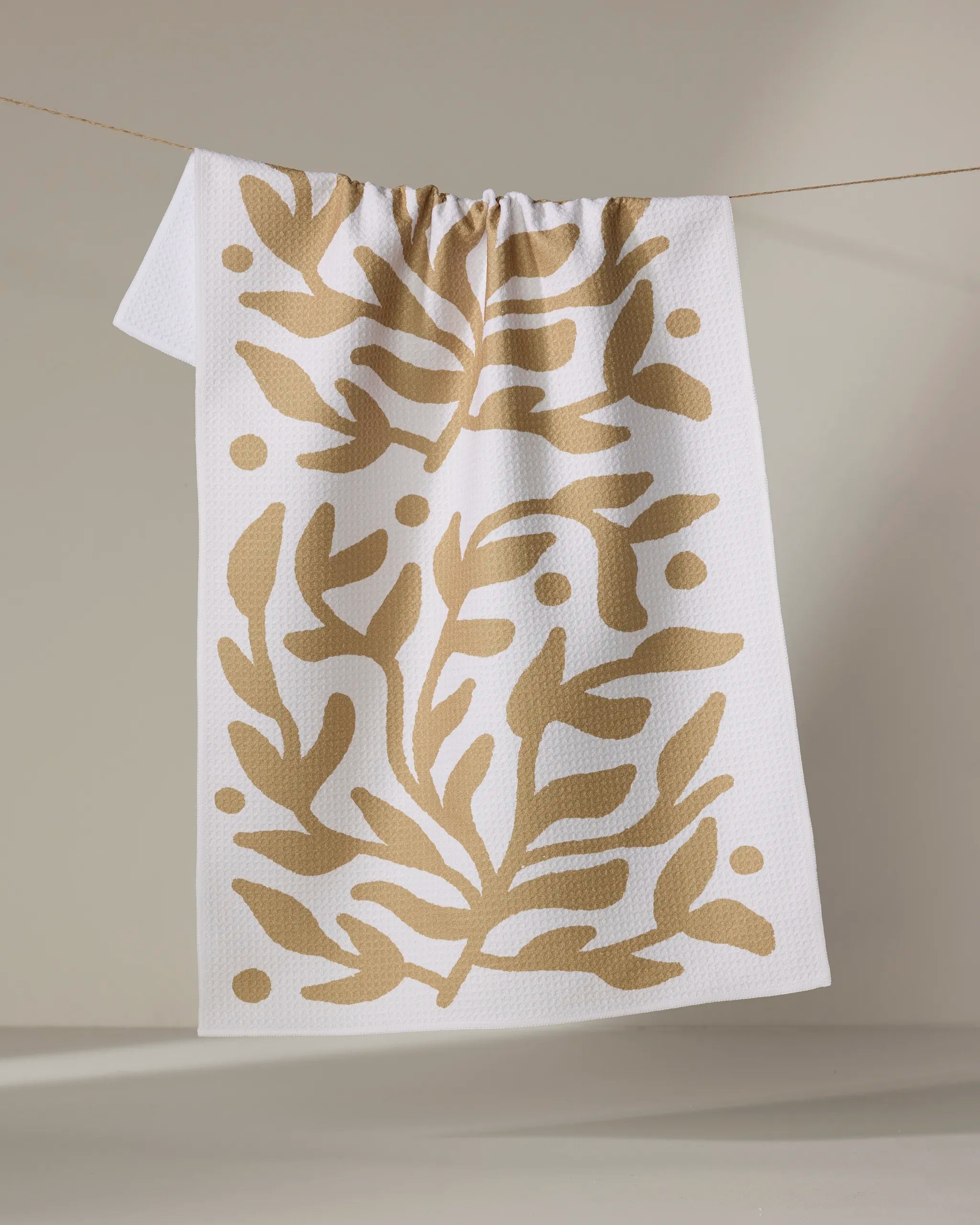 The Golden Garden towel by Graphics and Grain is ultra-absorbent with a white textured finish, featuring an abstract beige leaf and dot pattern. It hangs on a string against a neutral background.
