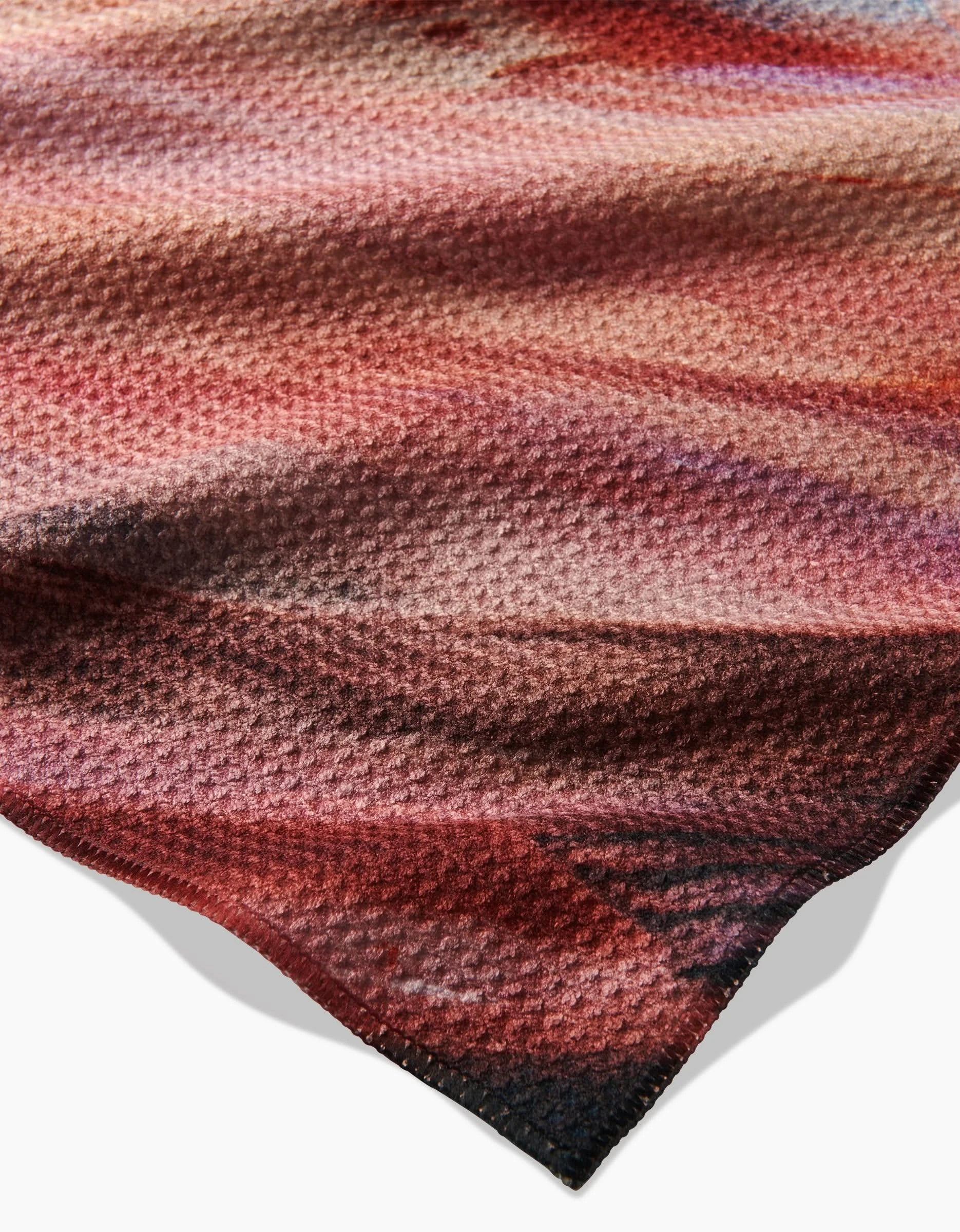 A close-up of the Golden Hour in the Rockies by Kelly Peters shows its textured, multicolored Geoweave fabric with wavy patterns and hues of red, pink, brown, and beige, featuring slightly uneven edges on a white background.