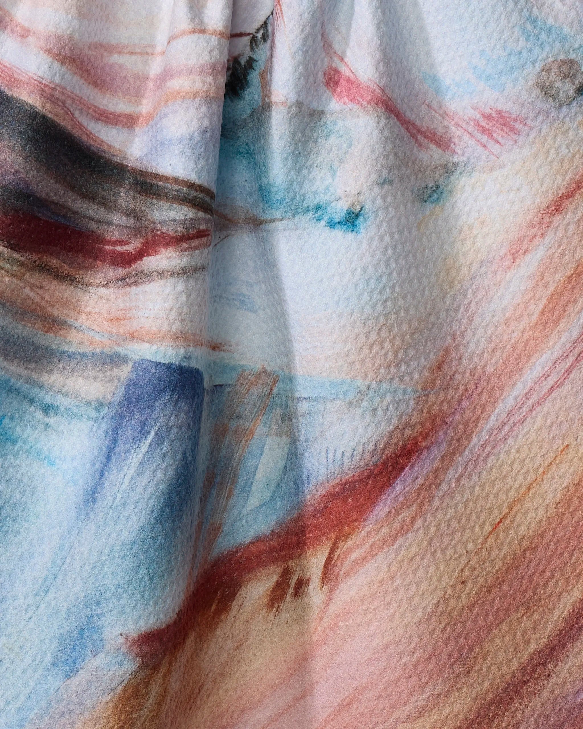 Close-up of Kelly Peters' "Golden Hour in the Rockies" tea towel, made from recycled materials, with soft watercolor-like streaks in blue, pink, brown, and red tones, gentle folds, and subtle shadows.
